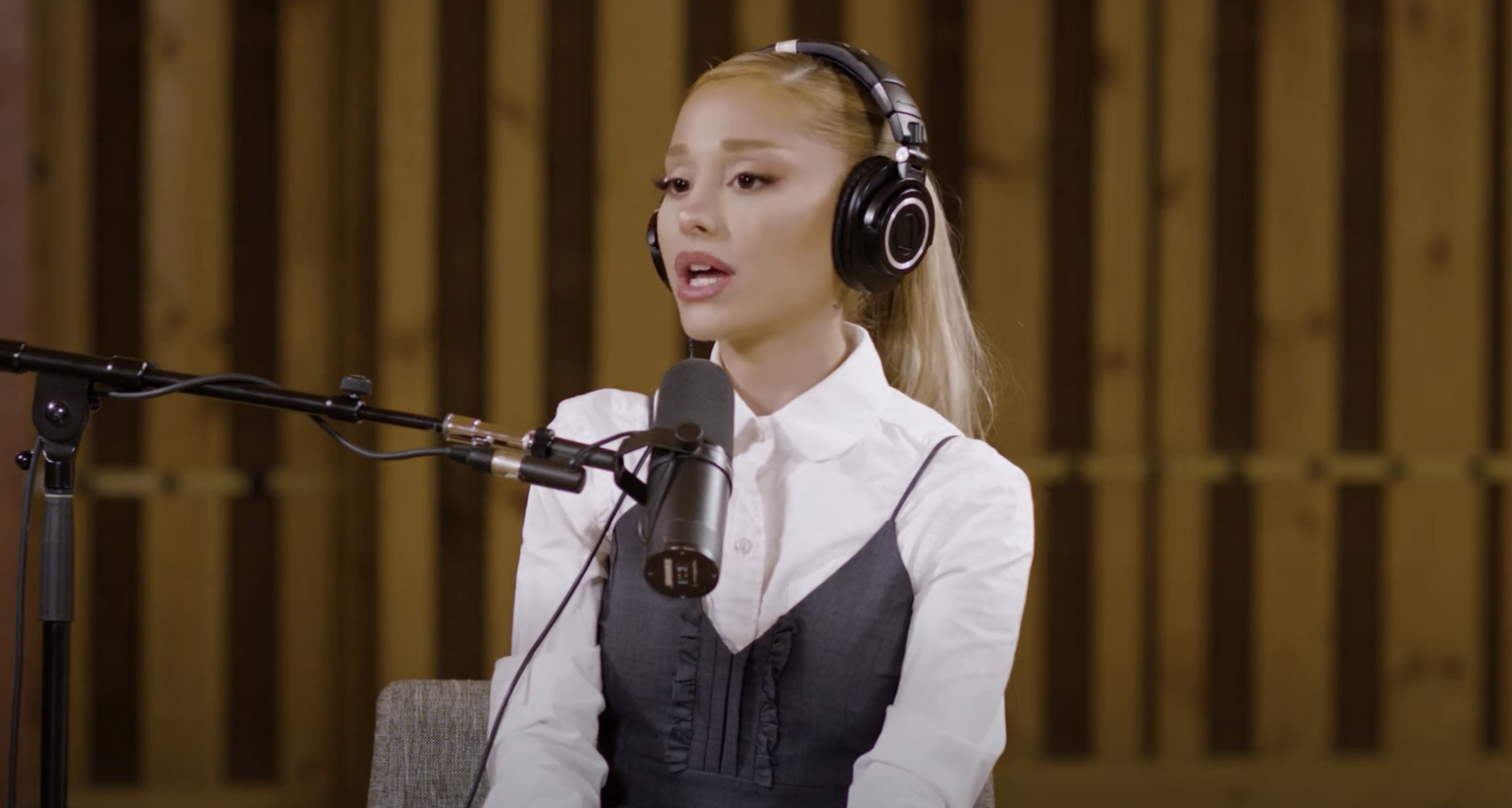 Ariana Grande speaking into a microphone in a recording studio, wearing headphones, a white shirt, and a grey vest