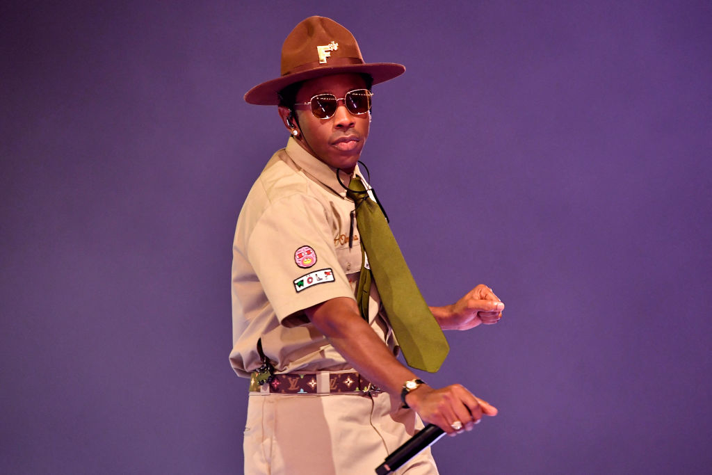 Tyler, the Creator, dressed in a beige scout uniform with patches, a wide-brimmed hat, sunglasses, and a green tie, performs on stage holding a microphone