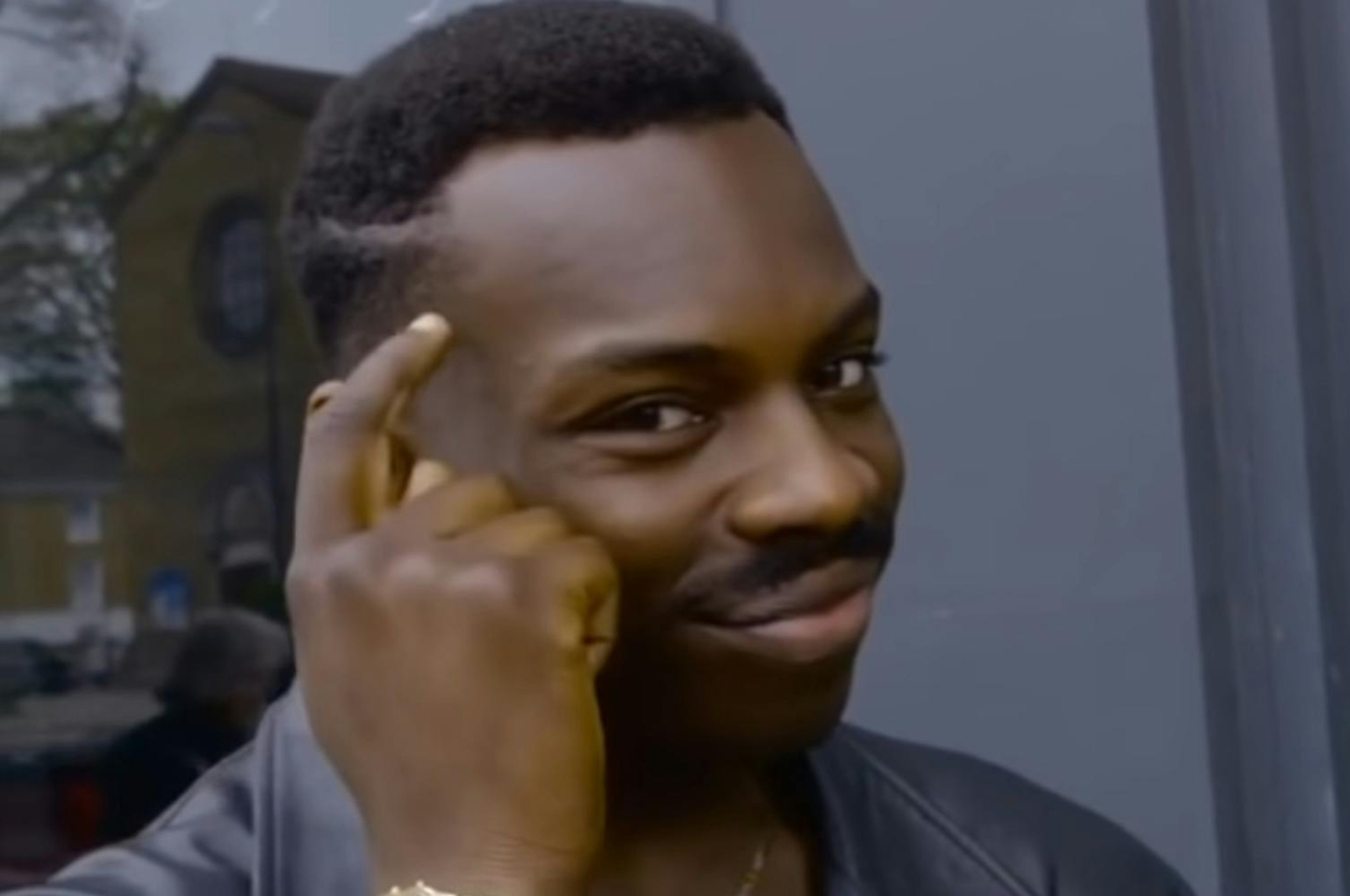 Man with short hair and mustache, wearing a leather jacket, taps his head with one finger near a window