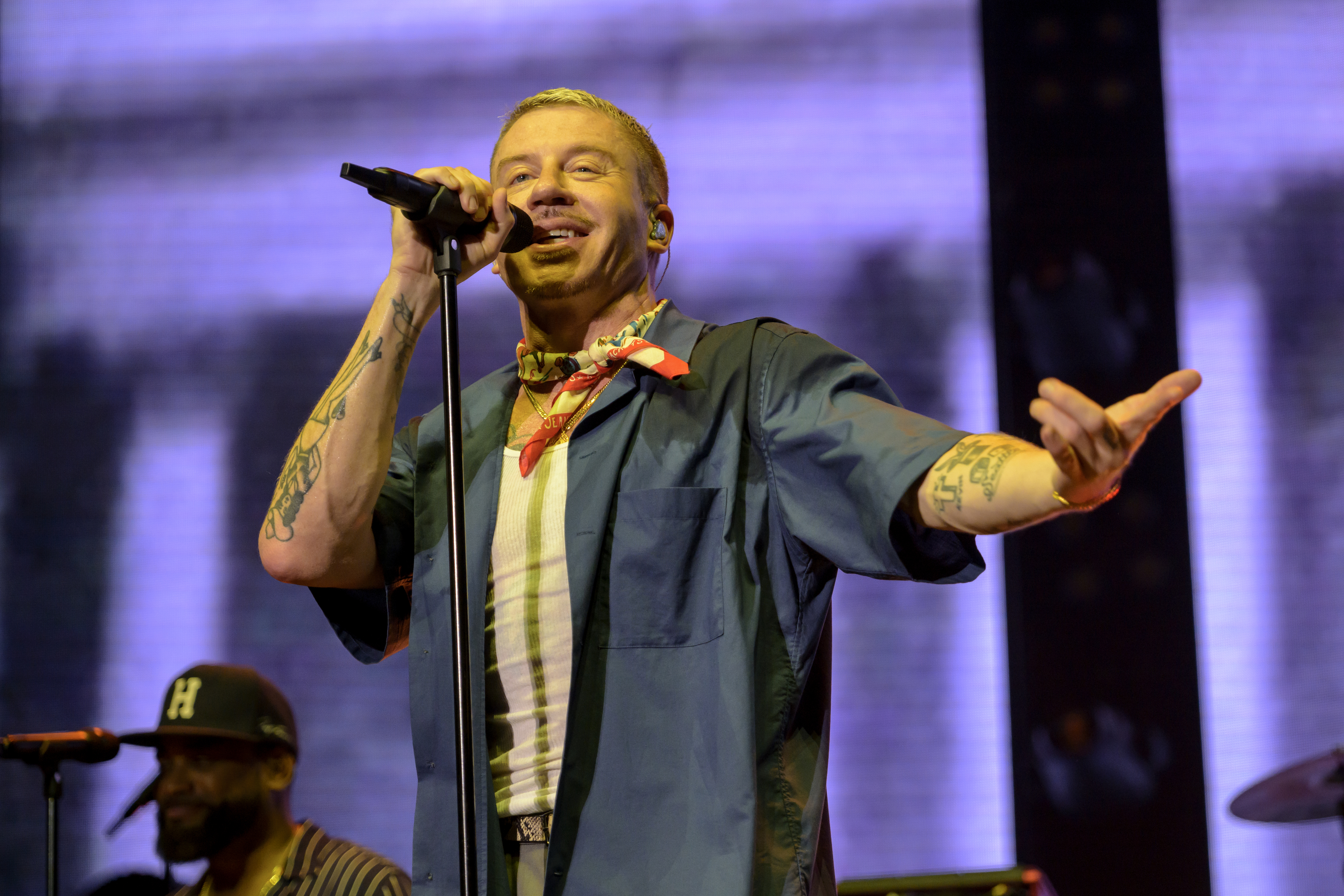 Singer Macklemore performs on stage, wearing a short-sleeved open jacket over a striped shirt and a bandana around his neck