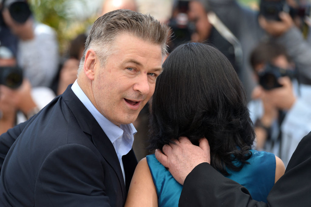 Alec Baldwin turns around, facing the camera with a smile, while standing next to a woman with black hair. Photographers and cameras are in the background
