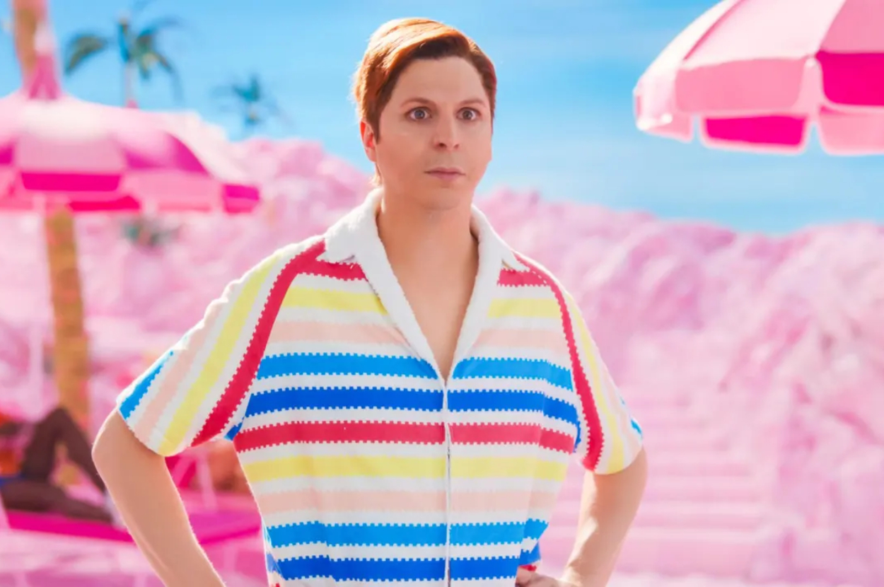Michael Cera stands on a beach set, wearing a striped short-sleeve shirt, with pink umbrellas and palm trees in the background