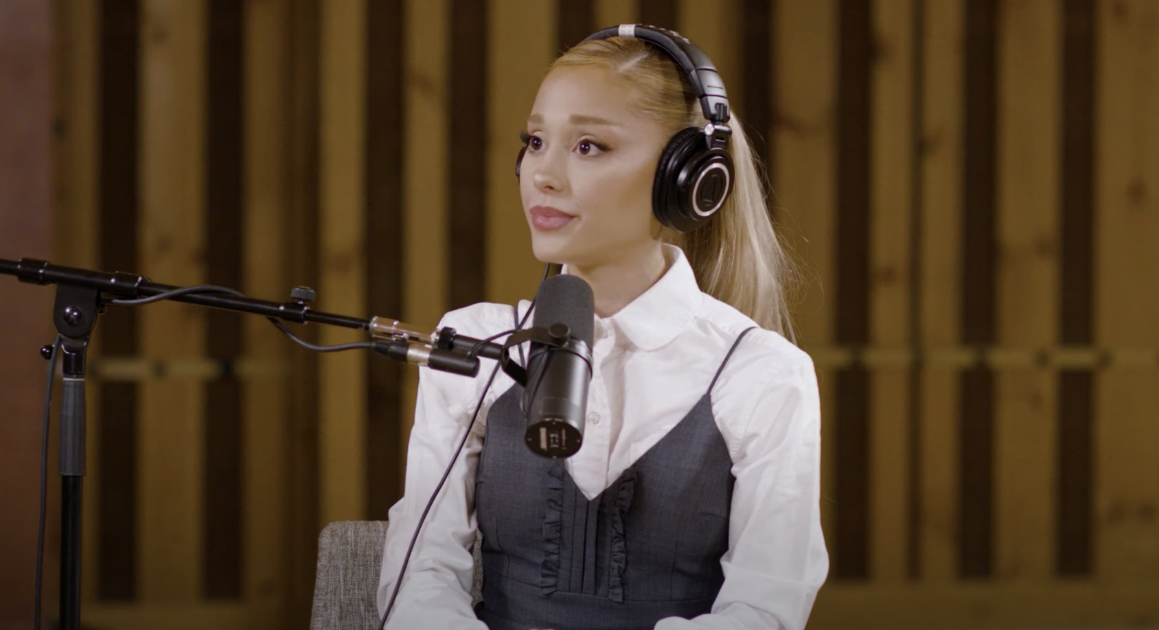 Ariana Grande speaks into a microphone during a podcast interview, wearing a blouse under a dress and headphones