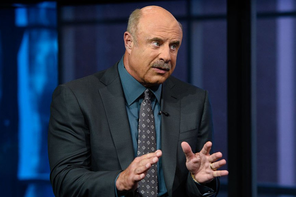 Dr. Phil is gesturing while speaking, dressed in a suit with a patterned tie, on a TV show set