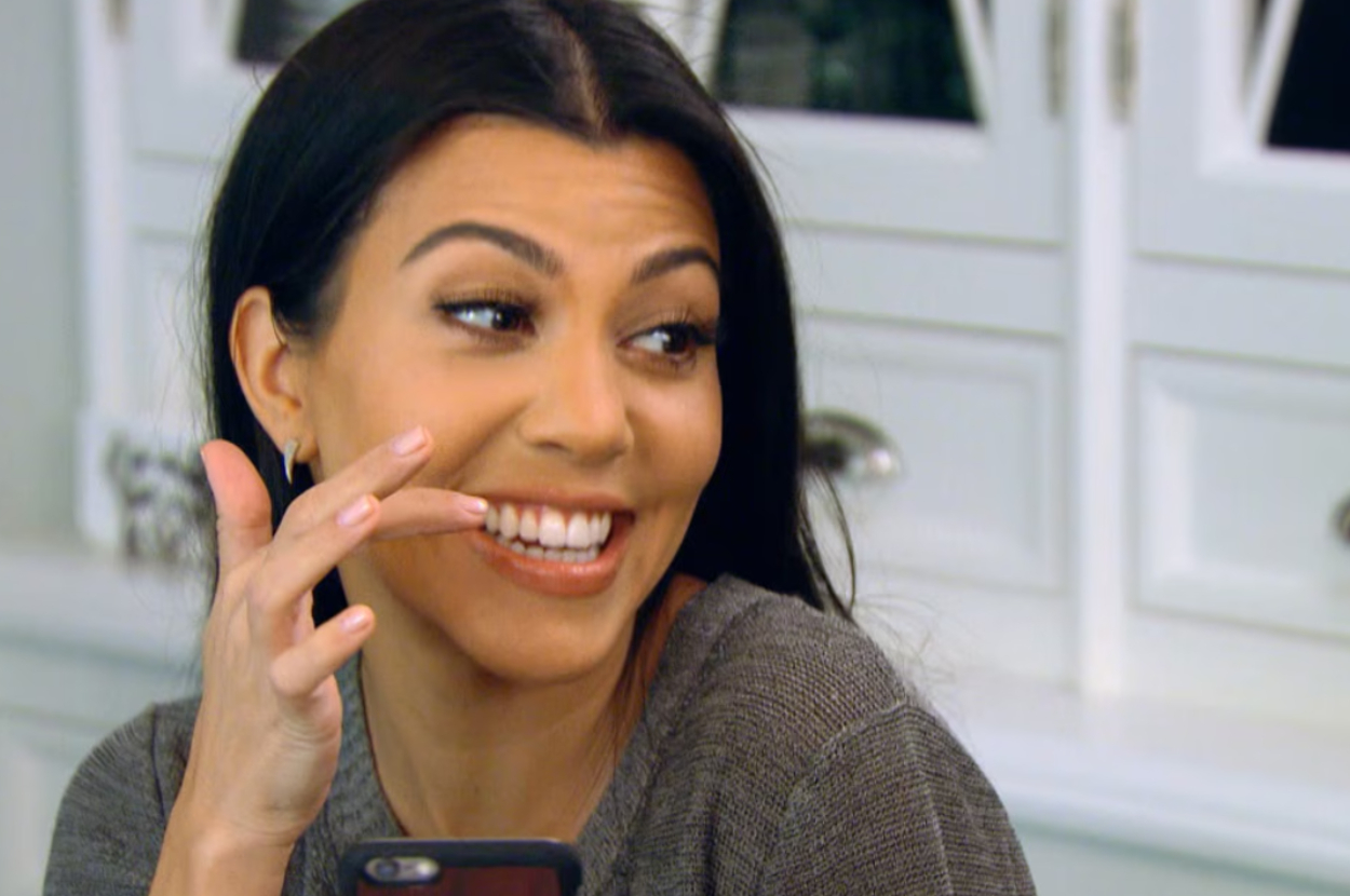 Kourtney Kardashian smiles and looks to the side while holding a phone. She has long, straight hair and is wearing a casual top