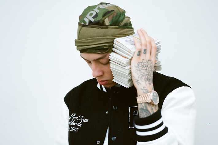 A person wearing a camo beanie and a black and white varsity jacket holds a large stack of money to their ear like a phone. Their hand has tattoos