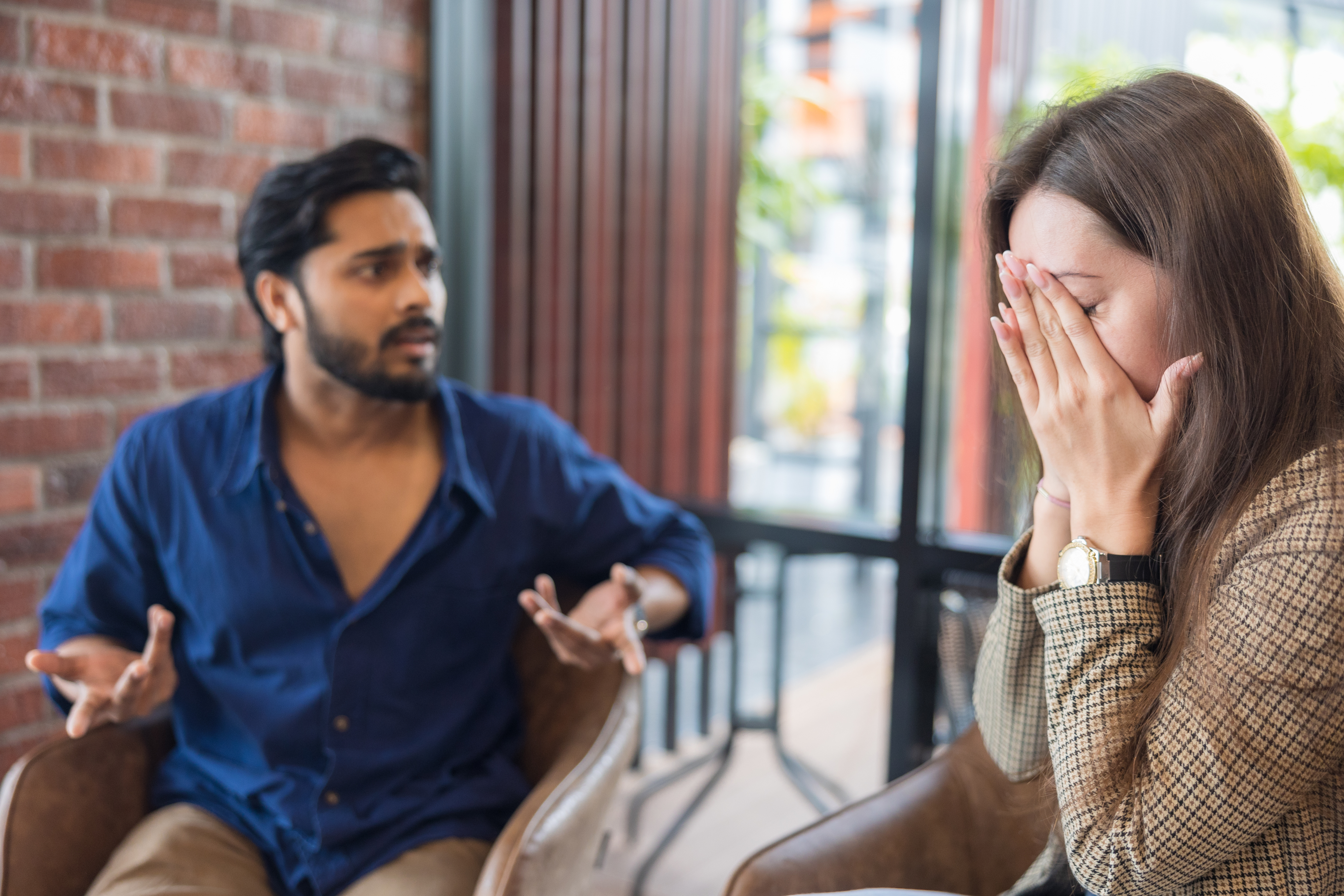 Man and woman in a tense conversation; woman covers face with hands, looking distressed, while man gestures emotionally