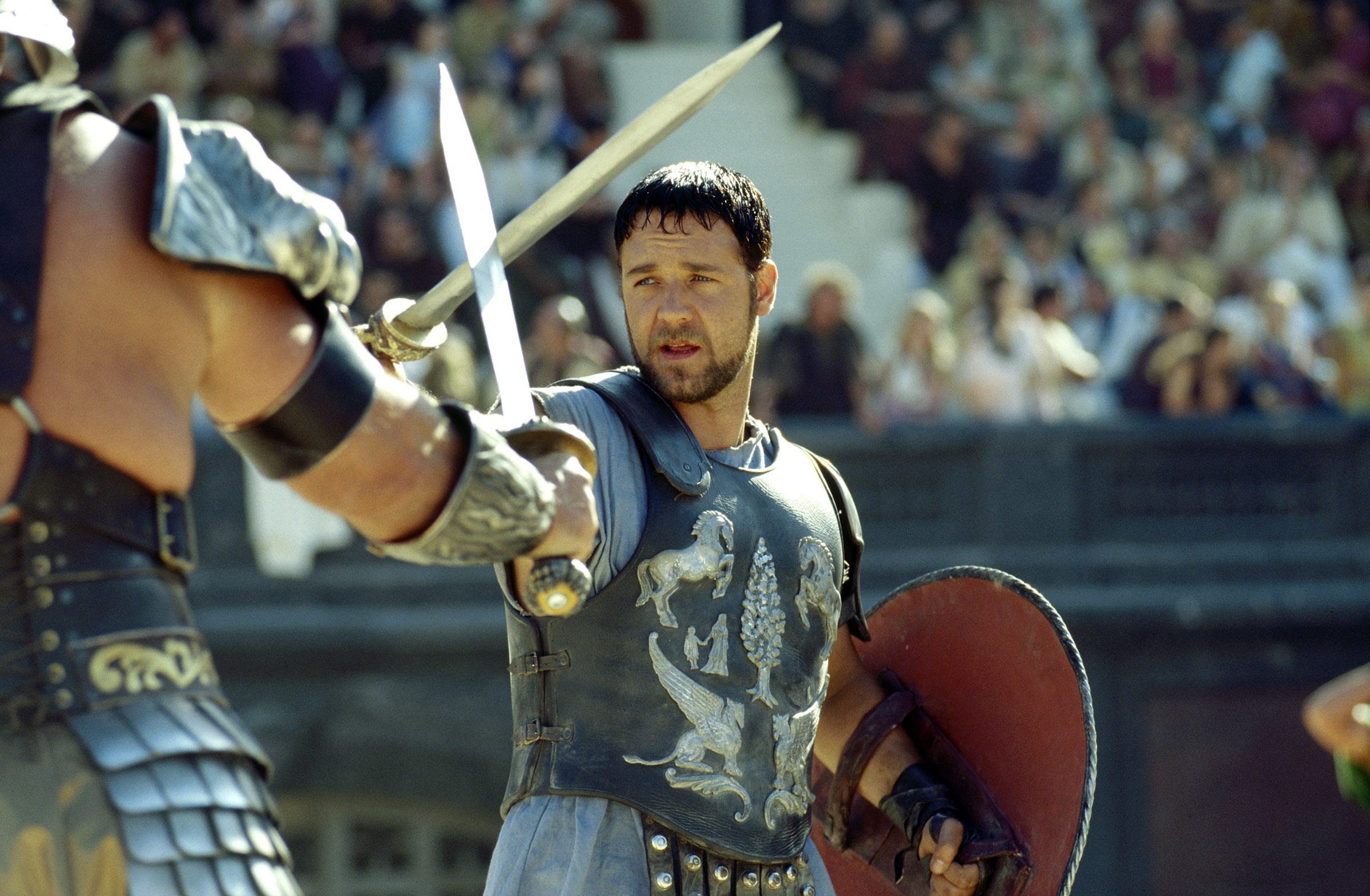 Russell Crowe dressed as a gladiator holds a sword and shield while facing another armored man with a sword