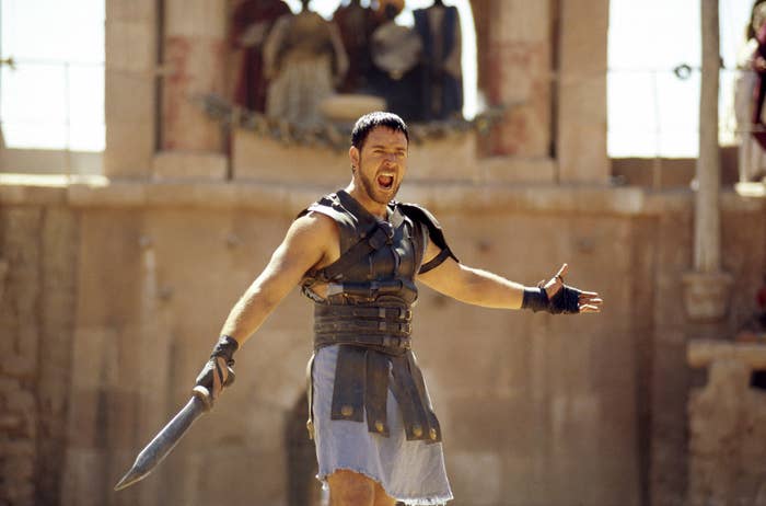 Russell Crowe, dressed as a Roman soldier with a sword and armor, stands in an ancient arena, appearing to shout with arms outstretched