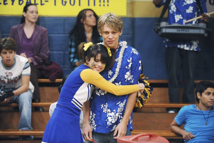 Selena Gomez hugs Austin Butler, both smiling. Selena is in a cheerleader outfit; Hutch wears a floral shirt. They are in a gym, crowd in the background