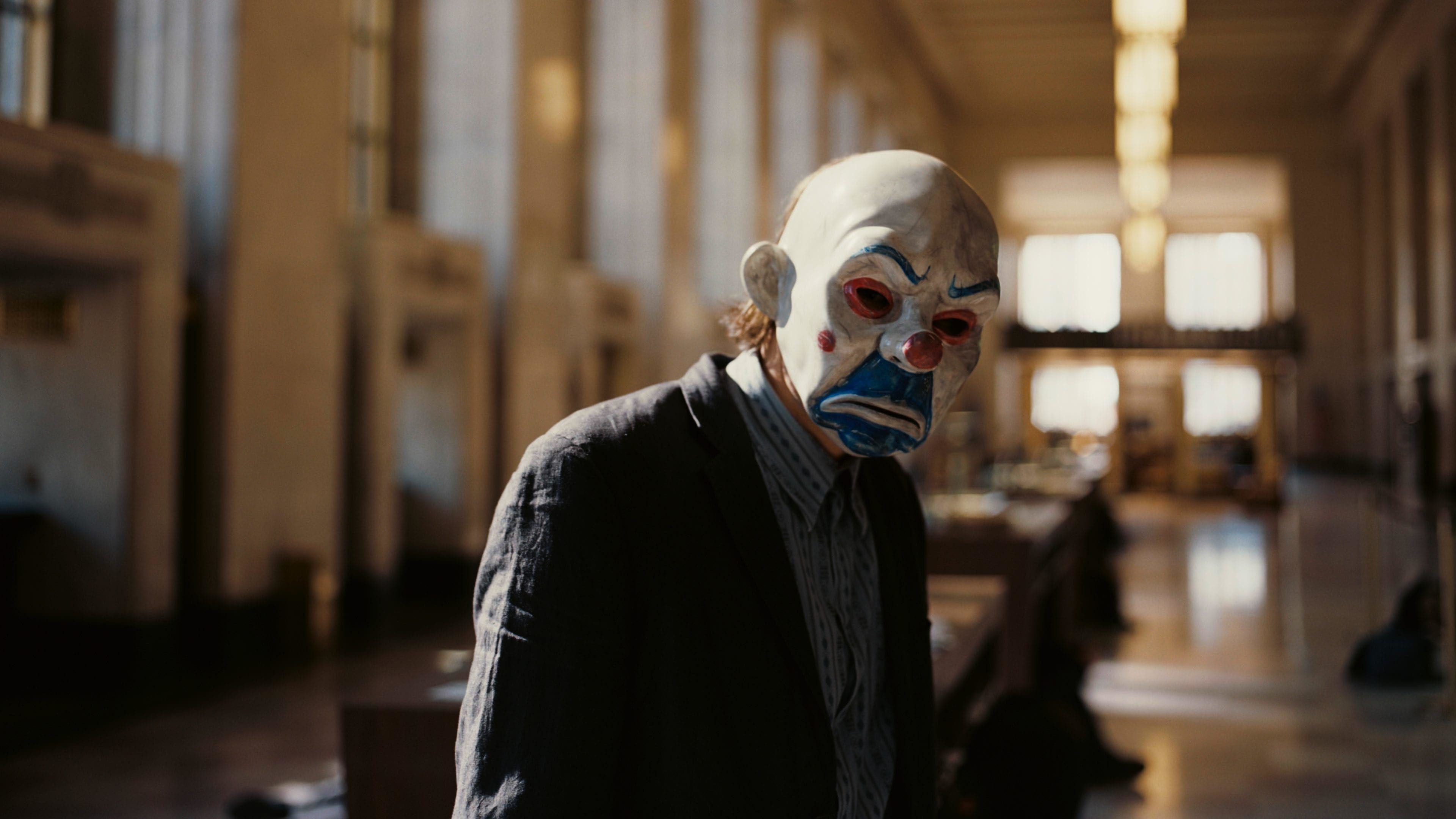A character in a worn suit and a clown mask with exaggerated features stands in a large, empty building with high ceilings and rows of windows