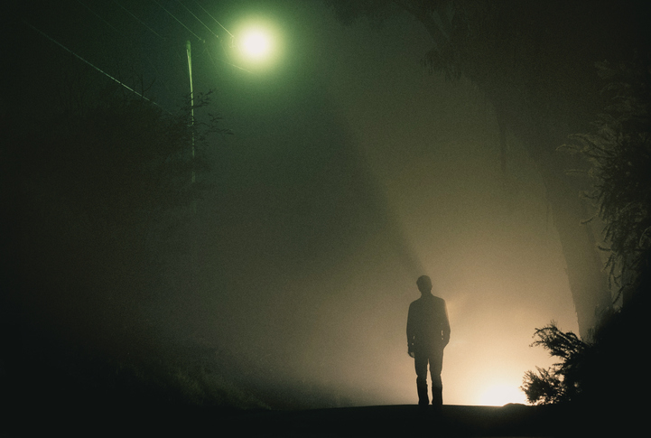 A silhouette of a person stands on a foggy road at night, illuminated by a streetlight and a distant light source behind them