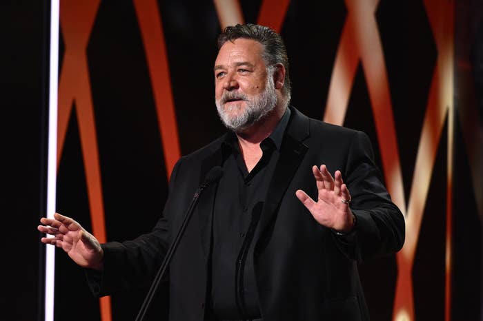 Russell Crowe in a black suit and black shirt, speaking on stage with hands slightly raised, red and orange background