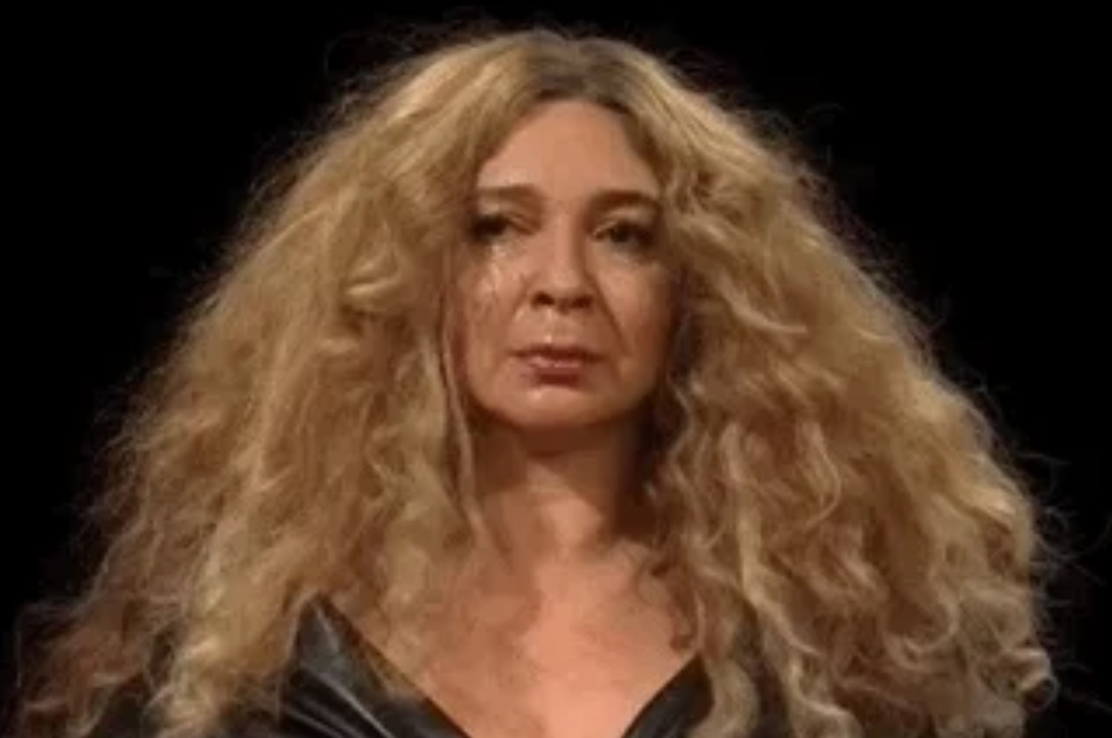 A woman with long, curly hair stands in front of a black background, wearing a shiny, dark-colored top
