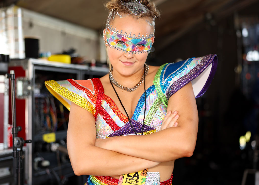 JoJo Siwa standing with arms crossed in an elaborate, sparkly outfit with rainbow accents and a glittery mask, smiling at LA Pride event