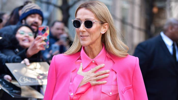 Celine Dion in a pink outfit with sunglasses, smiling and placing her hand on her chest as fans in the background take photos and hold up albums for autographs