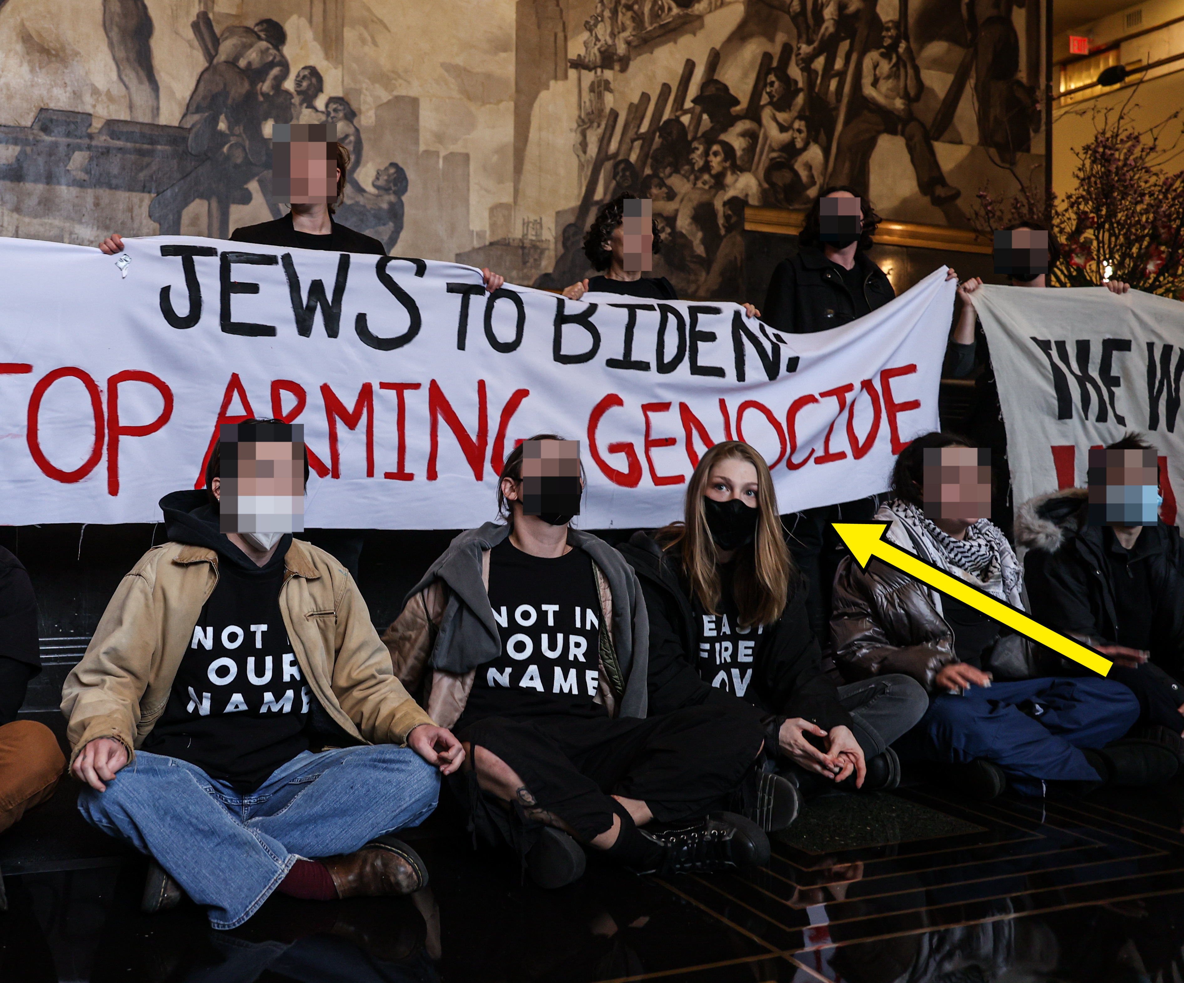 Group of protestors sitting and holding signs, including &quot;Jews to Biden: Stop Arming Genocide&quot; and wearing masks and &quot;Not In Our Name&quot; shirts
