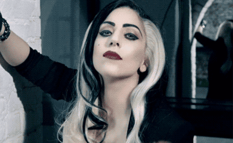 Lady Gaga posing with an arm on a wall, wearing bold makeup and an outfit giving a gothic vibe. Her two-toned hair is styled in loose waves