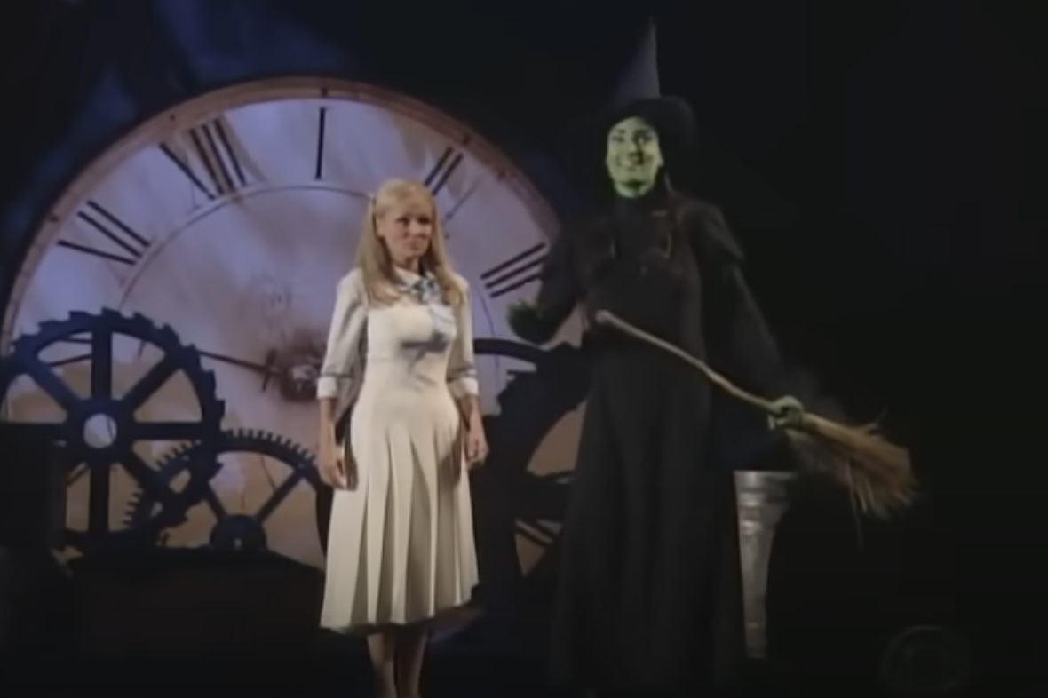 Kristin Chenoweth in a cream dress and Idina Menzel in a witch costume with green makeup stand in front of a large clock on stage