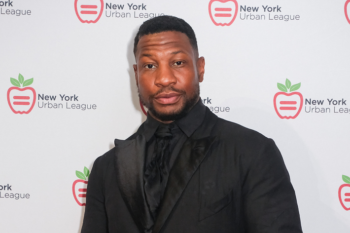 Jonathan Majors in a black suit and tie at the New York Urban League event, standing in front of a step and repeat backdrop with the event logo
