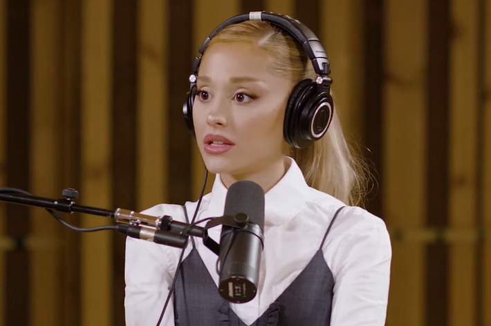 Ariana Grande wearing headphones and a white blouse layered with a dark vest, speaking into a microphone in a recording studio