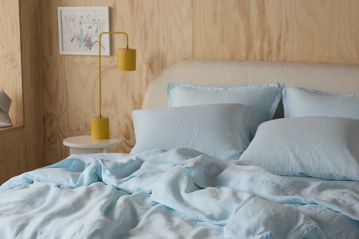 A neatly made bed with light blue sheets and pillows, a small white side table with two yellow lamps, and artwork hanging on a wooden wall in the background