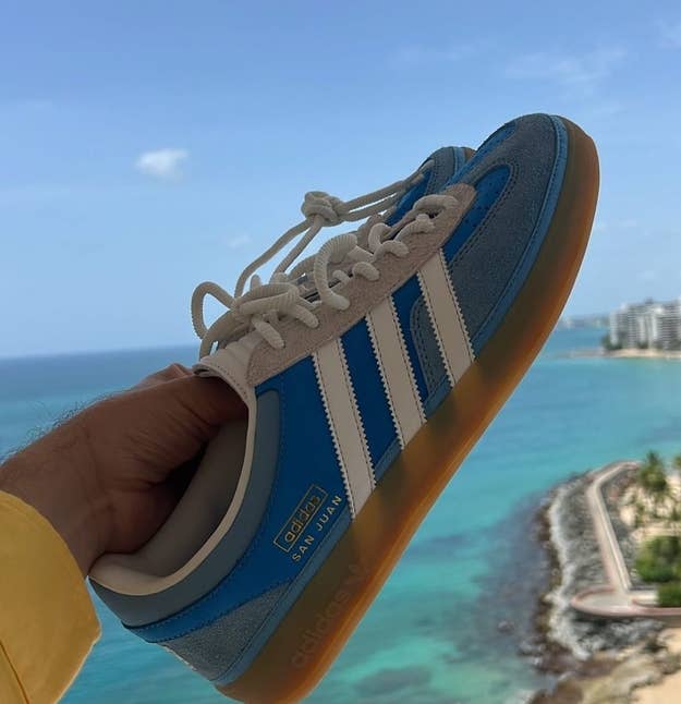A person's hand holds up a blue Adidas sneaker with white stripes and a golden sole, against a coastal ocean view