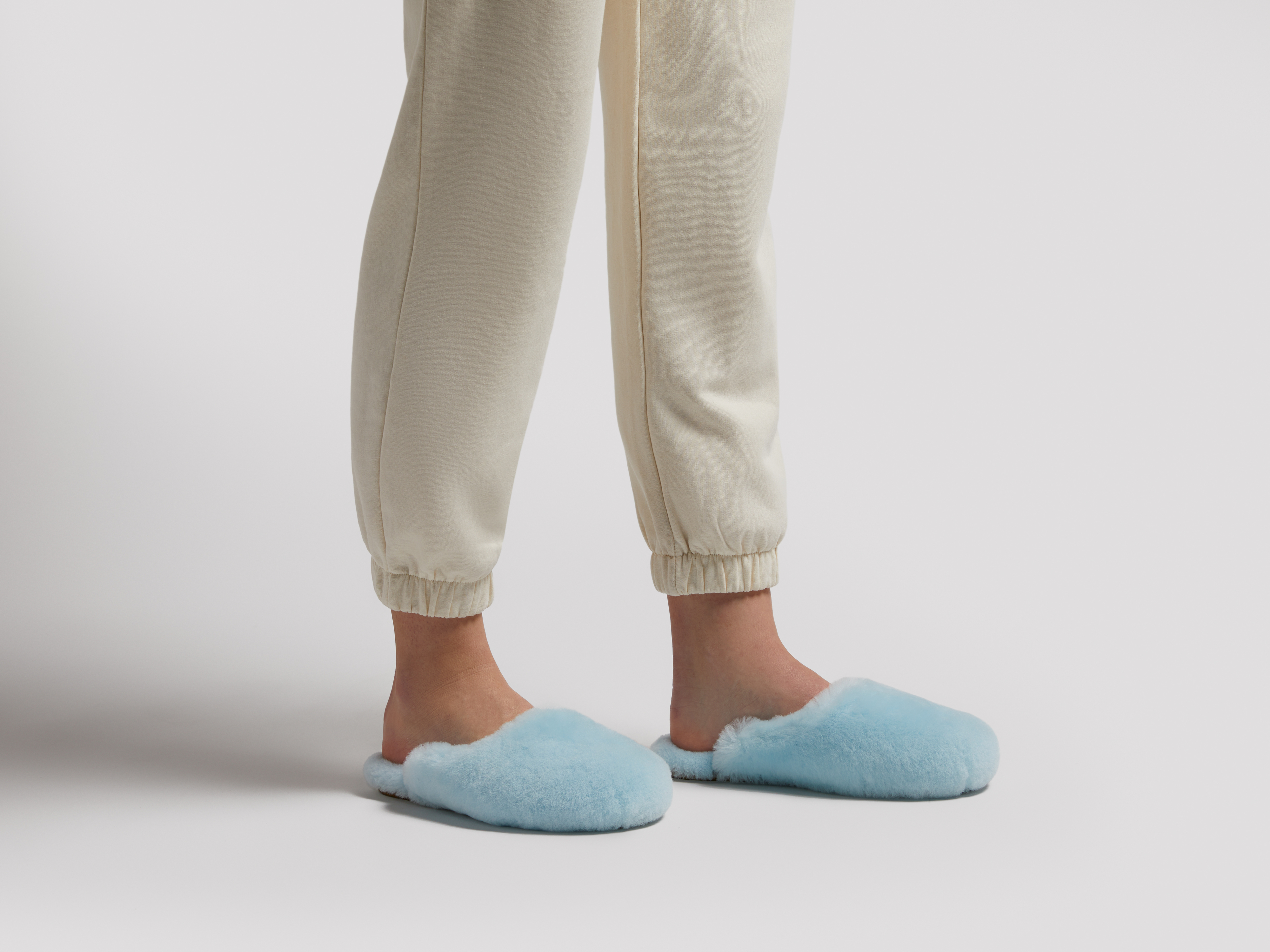 Feet wearing cream-colored sweatpants and fuzzy blue slippers, standing on a white background