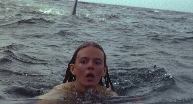 A woman looks terrified as she swims in the ocean, with a shark fin approaching in the background