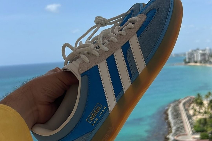 A person's hand holds up a blue Adidas sneaker with white stripes and a golden sole, against a coastal ocean view