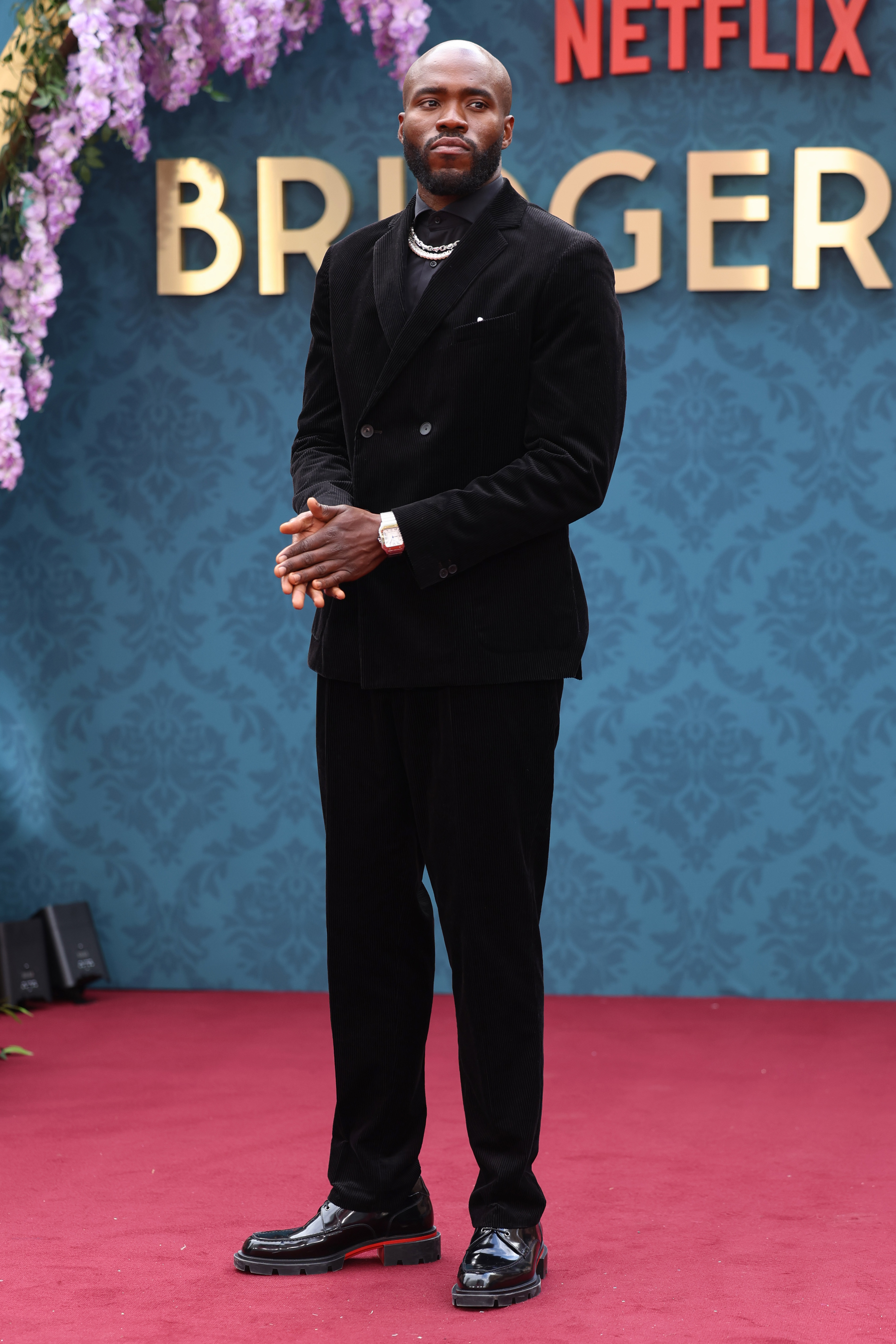 Martins Imhangbe dressed in an elegant black suit stands on a red carpet at a Netflix Bridgerton event backdrop