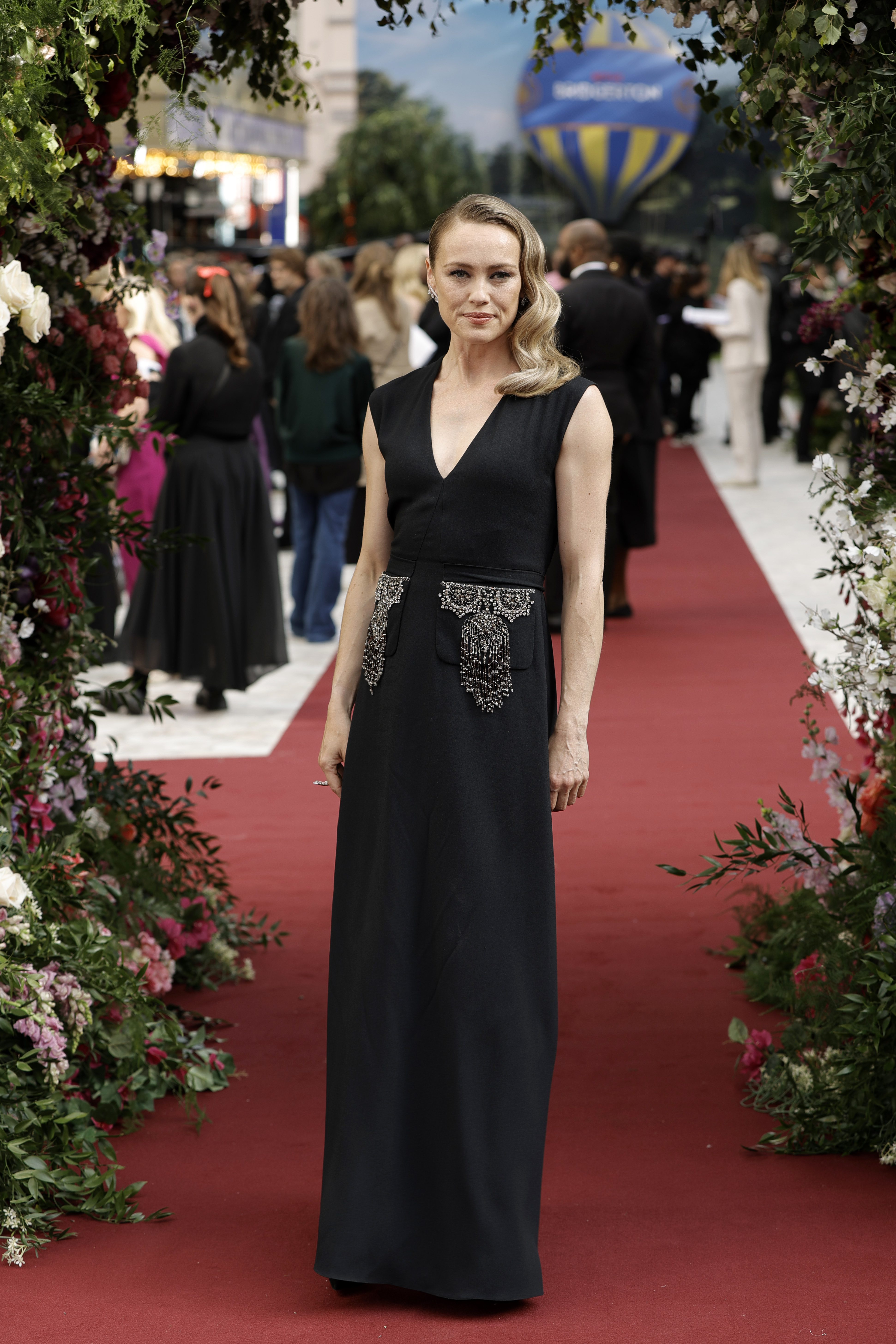 Hannah New in a sleek, floor-length black dress stands on a red carpet, surrounded by floral decorations. People and a hot air balloon are visible in the background