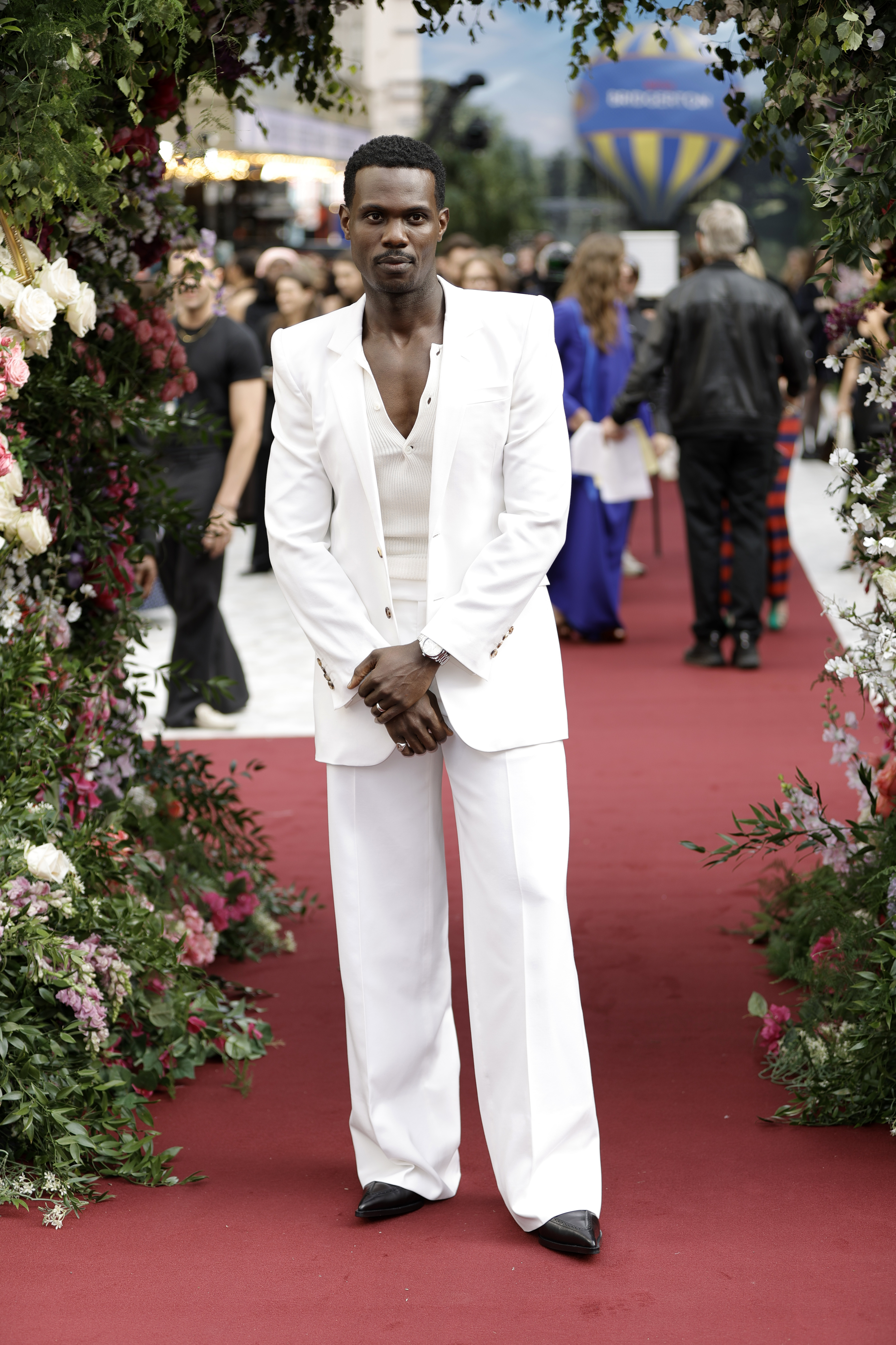 Victor Alli is on a red carpet wearing a white suit with a deep V-neck shirt, surrounded by lush floral arches and attendees in the background