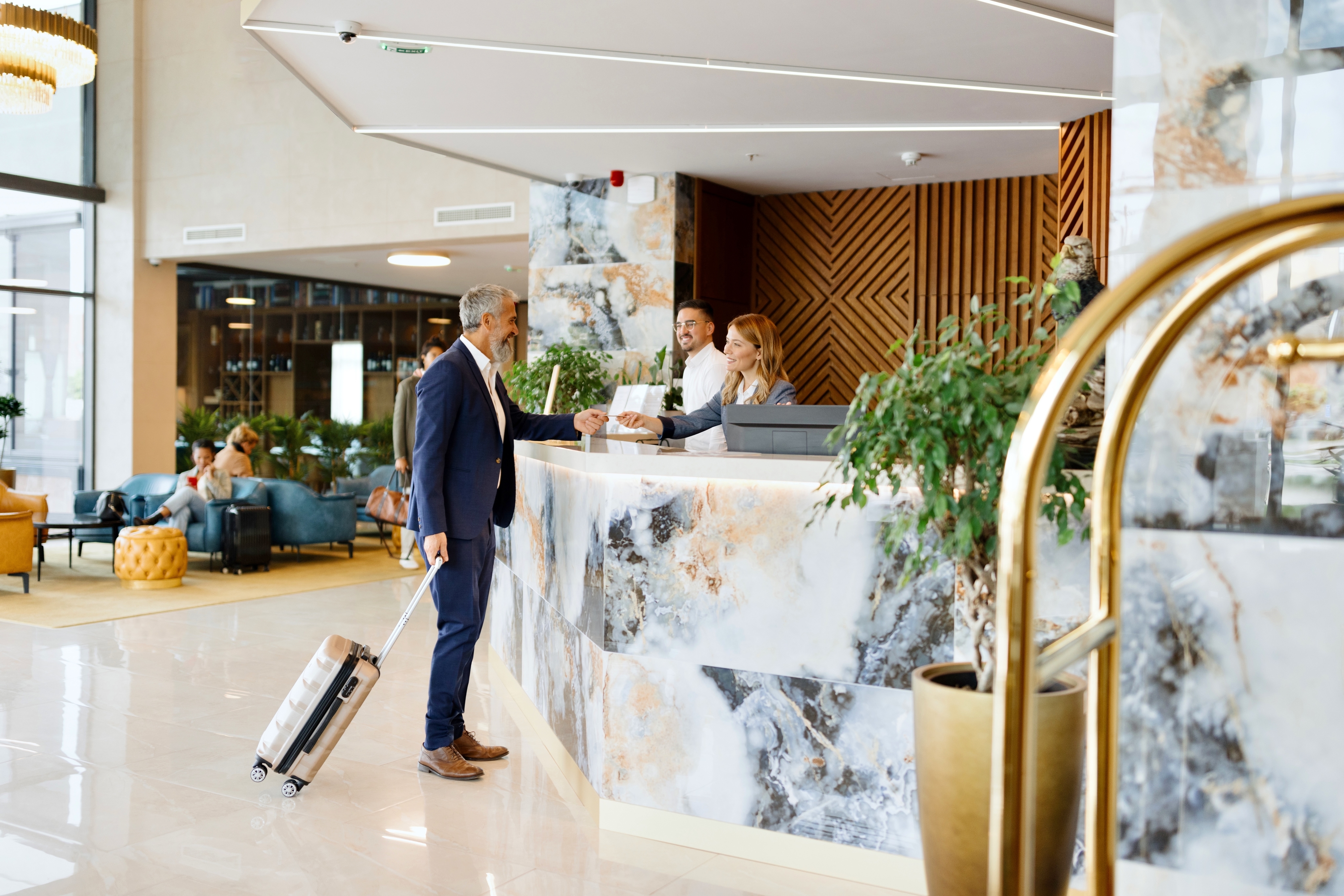 A man with grey hair and a suitcase checks in at a modern hotel reception desk attended by two staff members, one male and one female