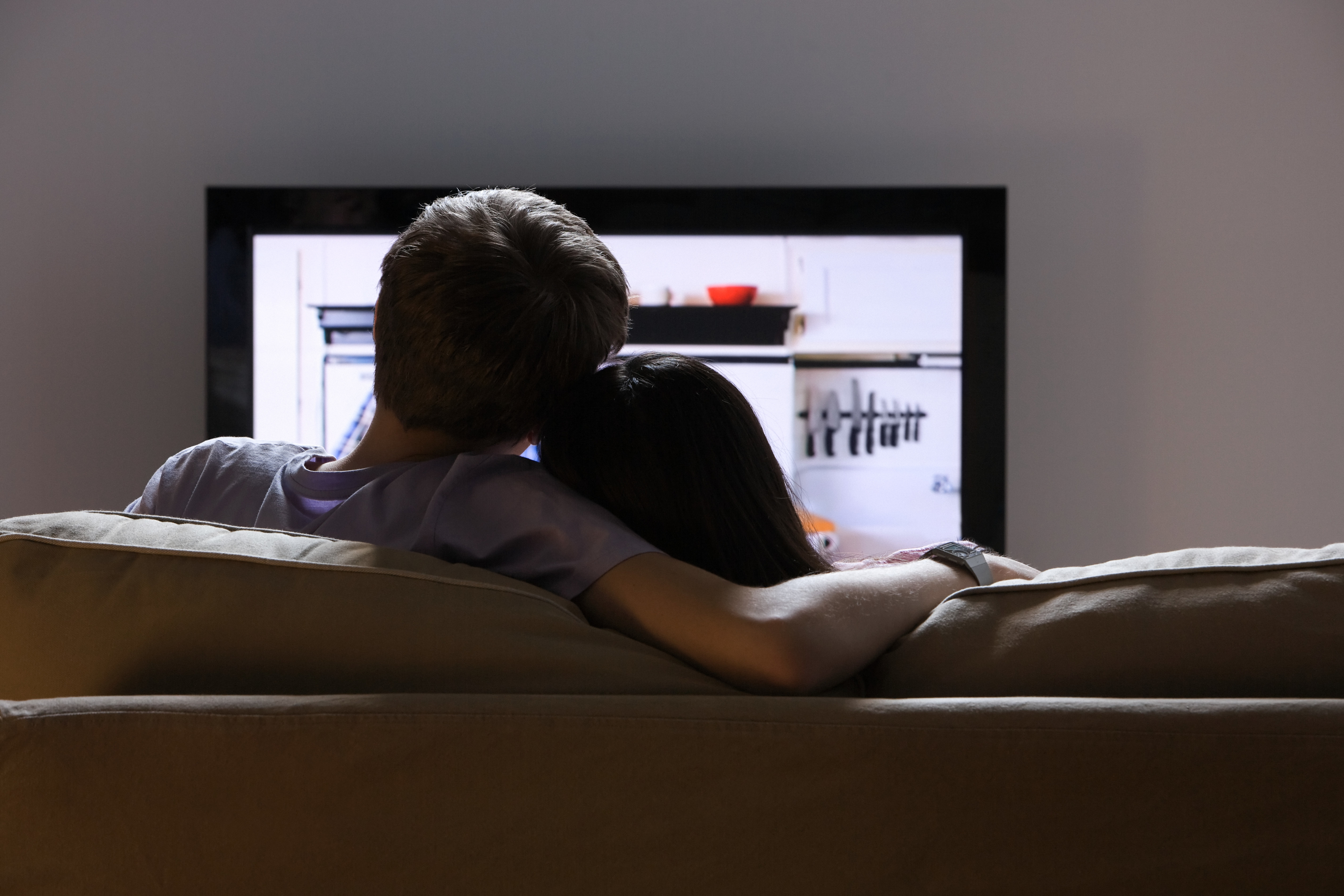 A couple sits on a couch with their backs facing the camera, watching TV together in a cozy setting