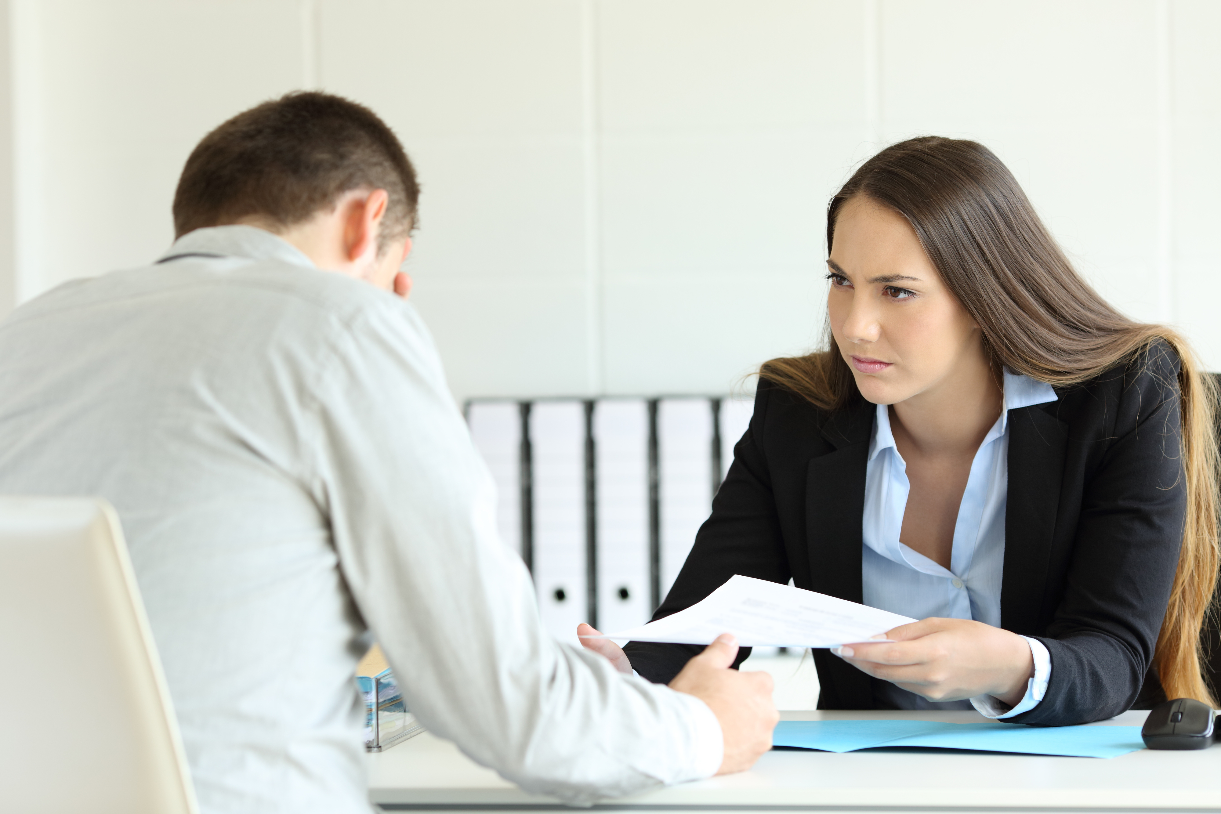 A woman in professional attire holds a document and talks with a man sitting across from her in an office setting