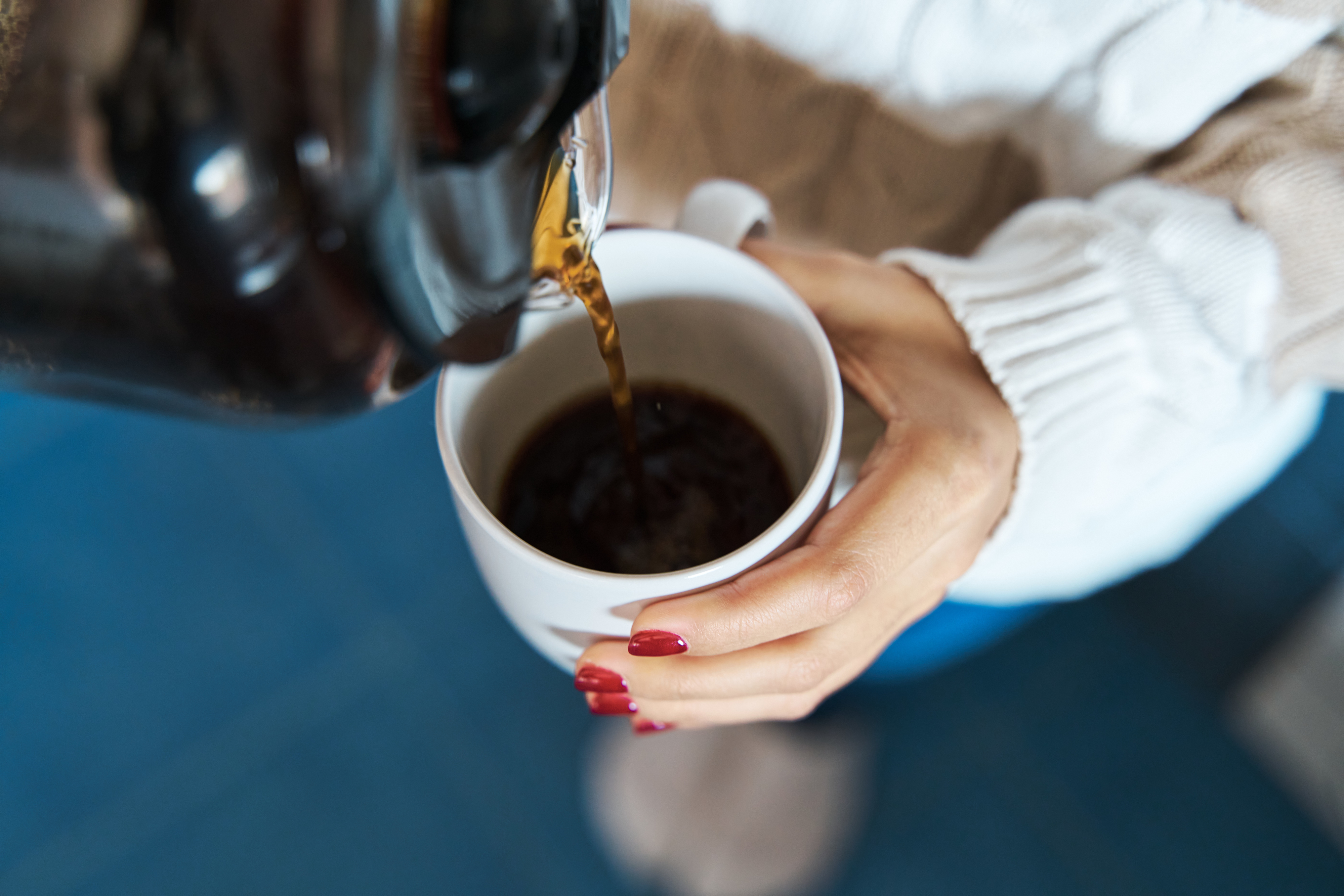 A person is pouring coffee from a pot into a white mug while holding the mug with the other hand. Their nails are painted, and they are wearing a sweater