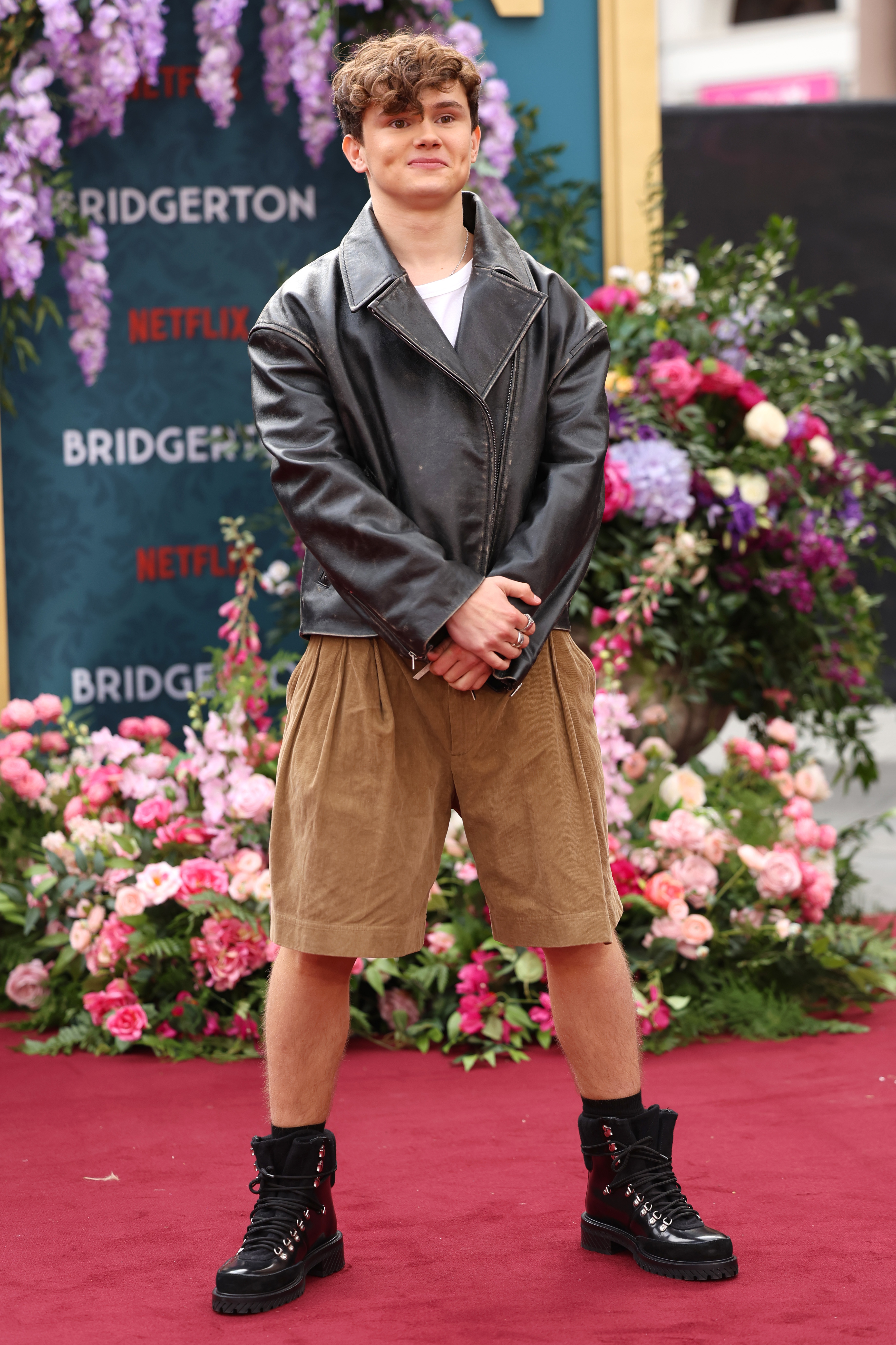 Will Tilston on a red carpet dressed in a leather jacket, khaki knee-length shorts, and black boots, posing in front of a floral backdrop at a Bridgerton Netflix event