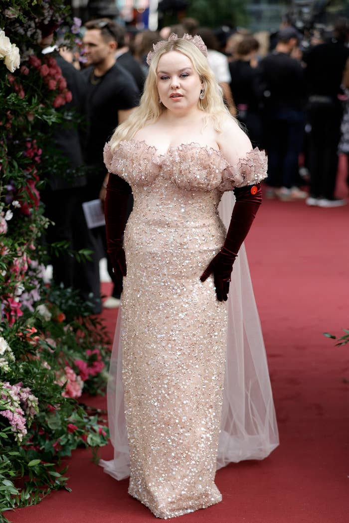 Nicola Coughlan on a red carpet, wearing an off-the-shoulder sequined gown with velvet gloves and a headpiece. Floral background and people in the distance