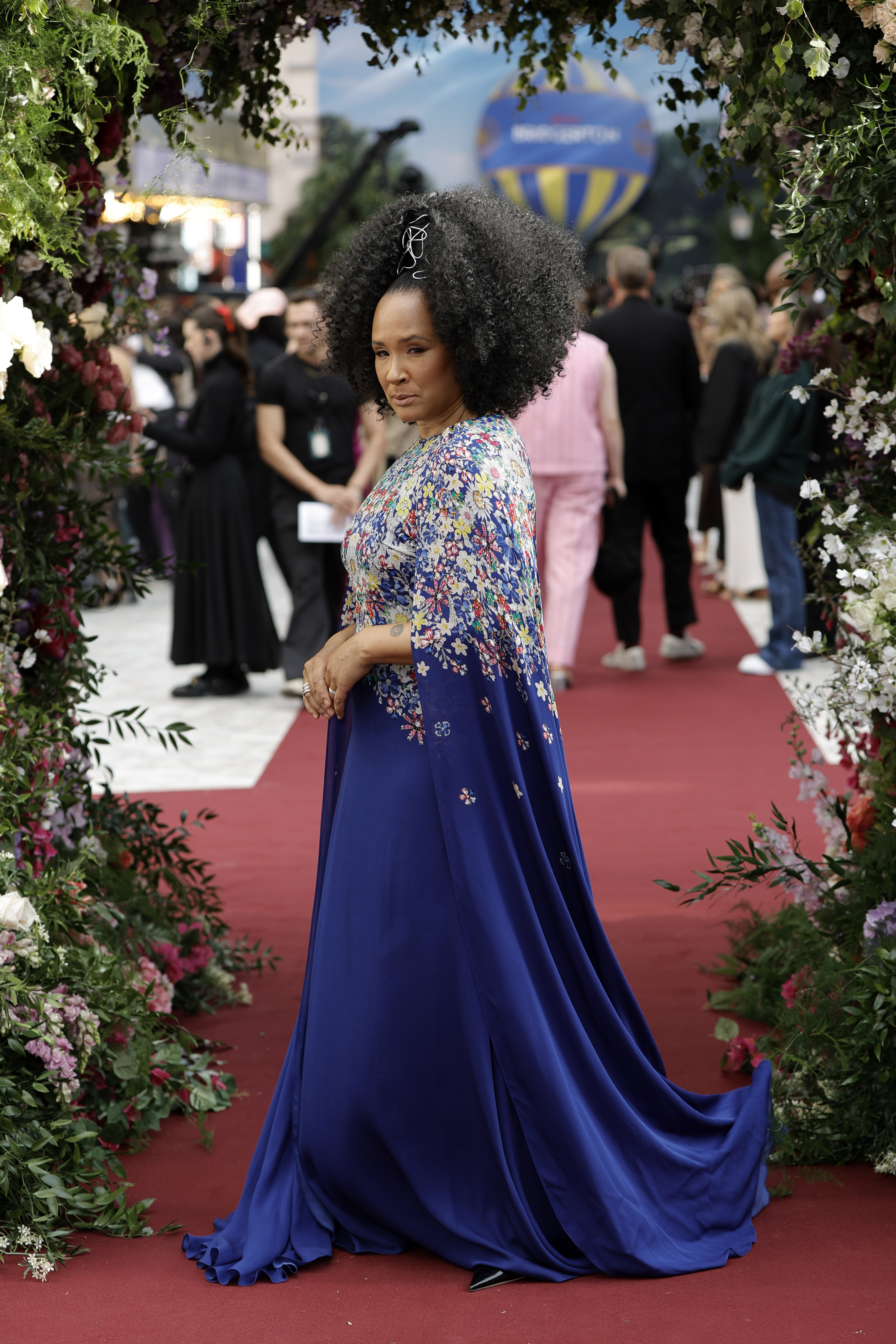 Golda Rosheuvel poses on the red carpet in a vibrant blue gown adorned with a floral cape, surrounded by floral decorations