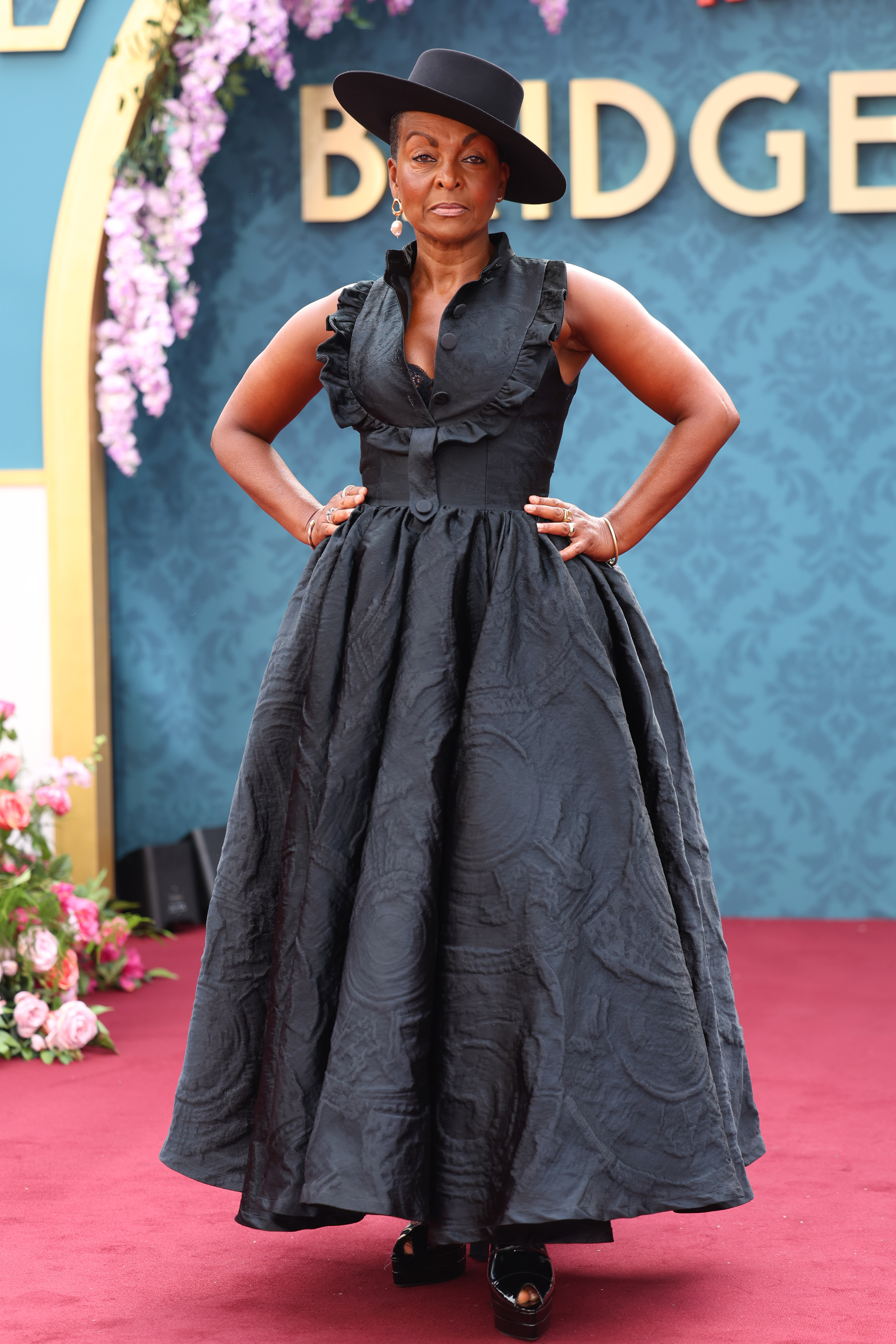 Adjoa Andoh stands on the red carpet wearing a black sleeveless gown and a wide-brimmed hat, accessorized with earrings and bracelets
