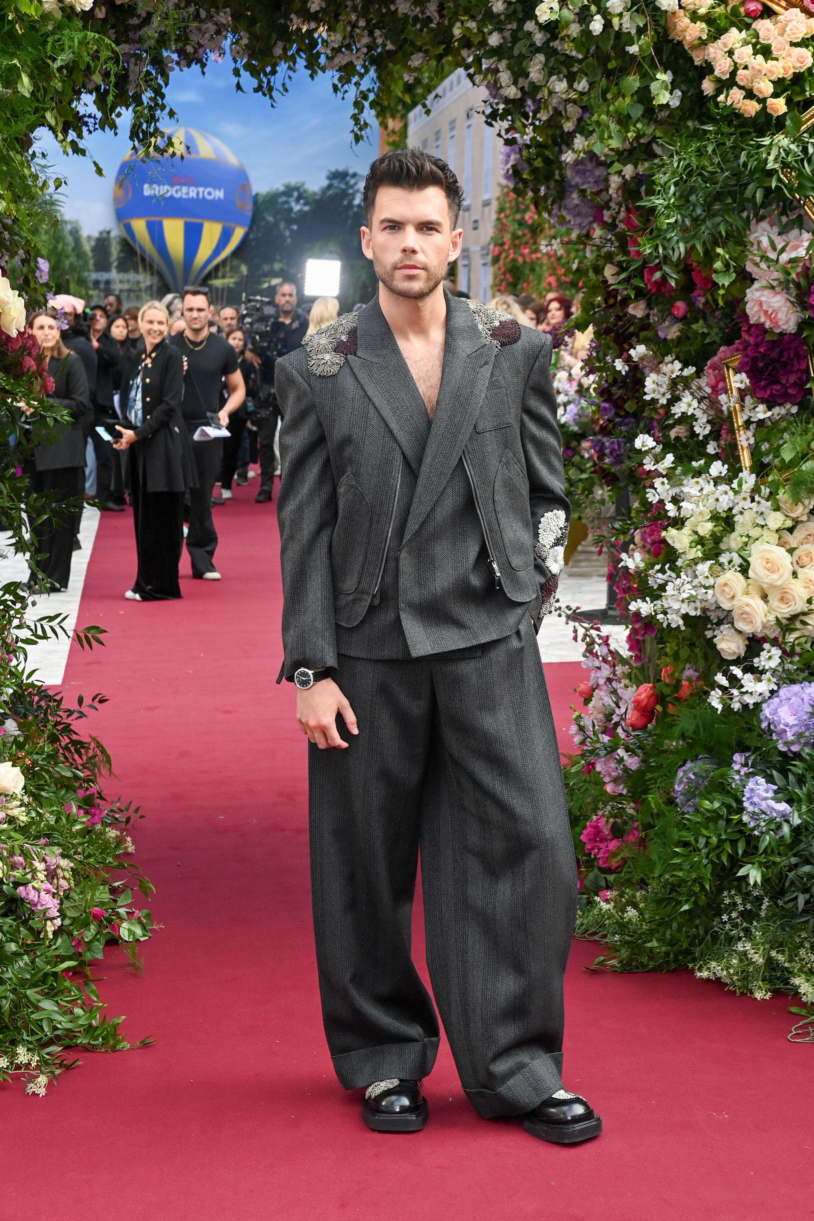 Luke Newton in a stylish, modern gray suit poses on a red carpet event under a floral arch, with a hot air balloon in the background