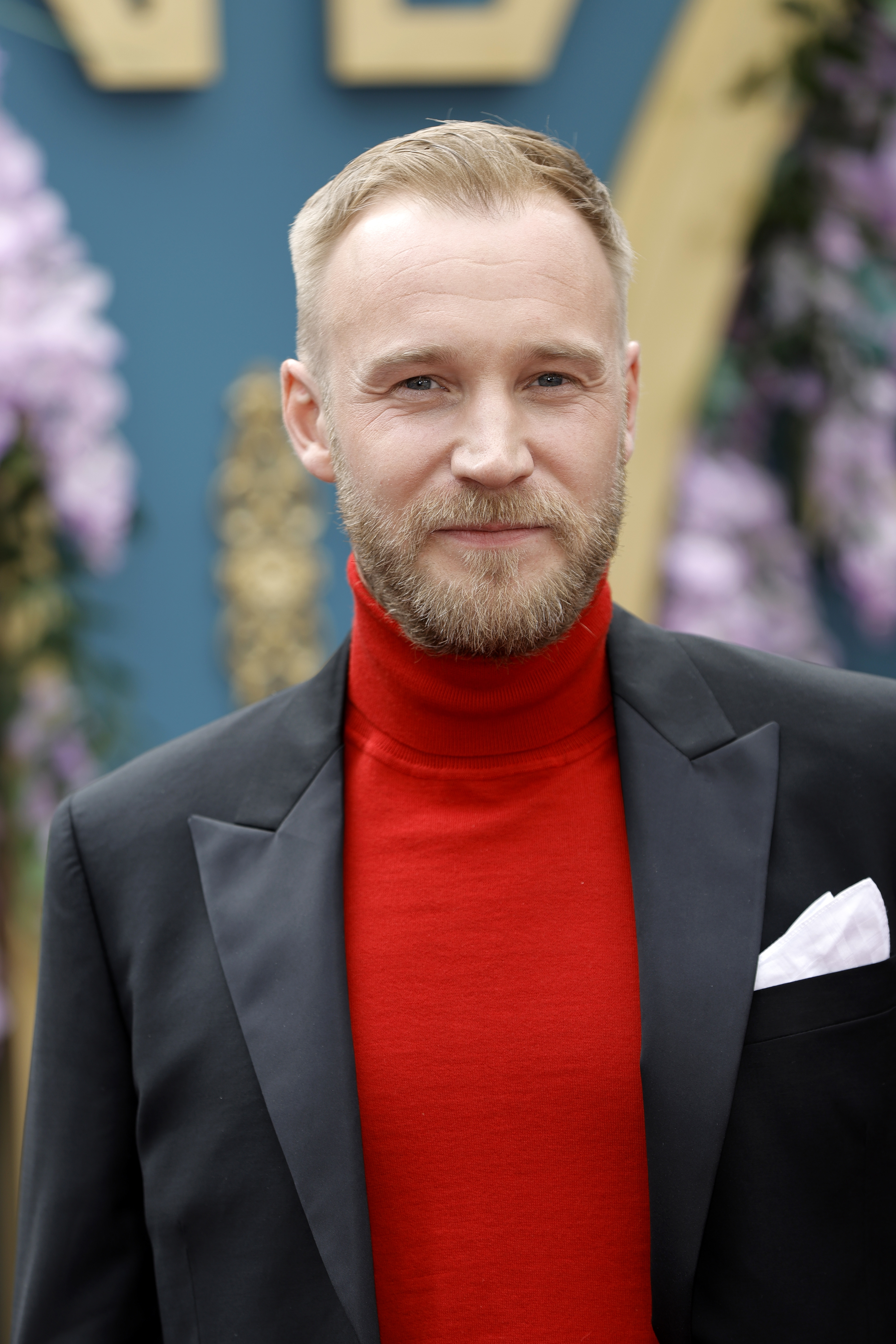 Sam Phillips in a red turtleneck and black blazer with a white pocket square on a red carpet