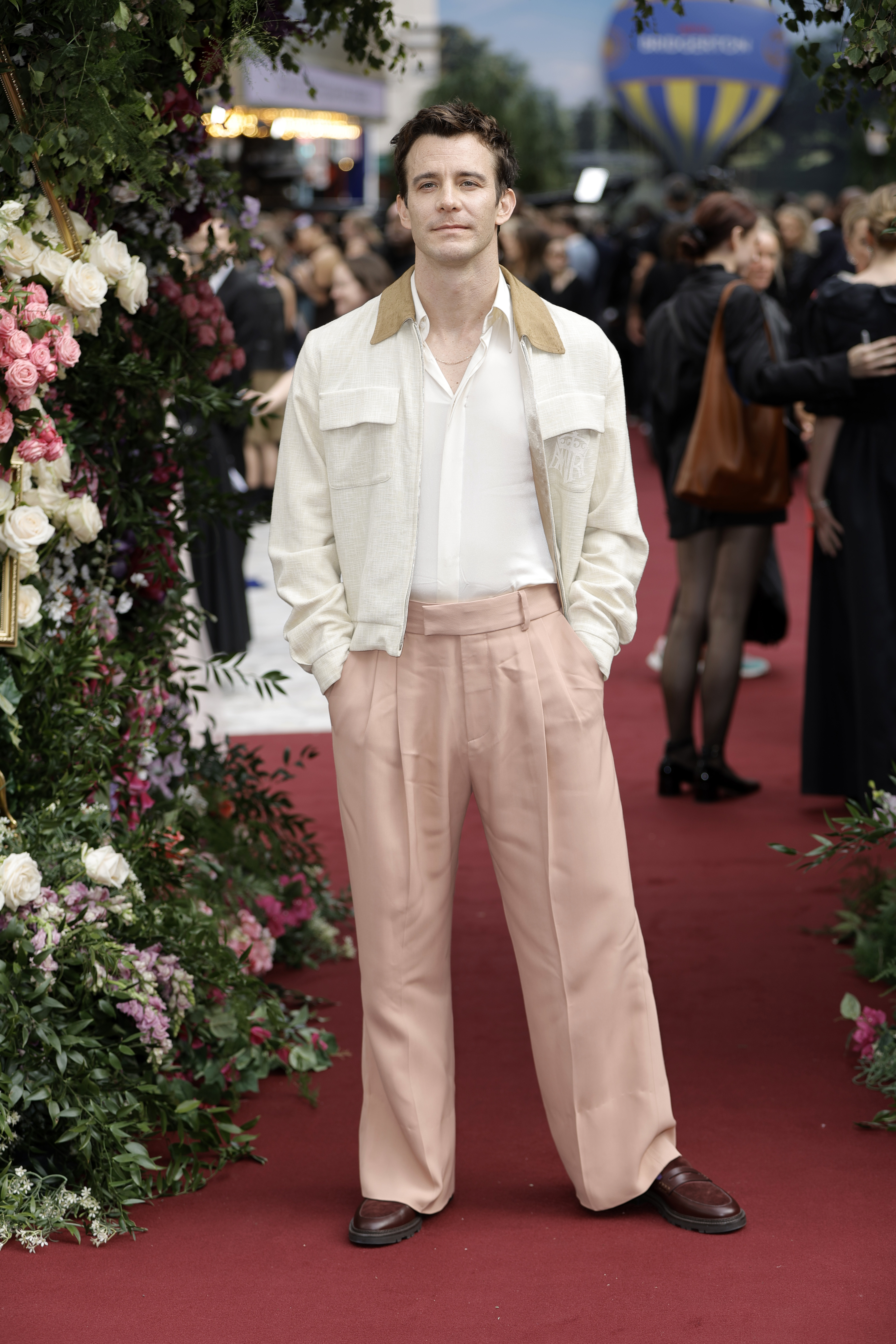 Luke Thompson stands on a red carpet, wearing a white shirt, beige jacket, and peach trousers, with hands in pockets. Floral decor and people are in the background