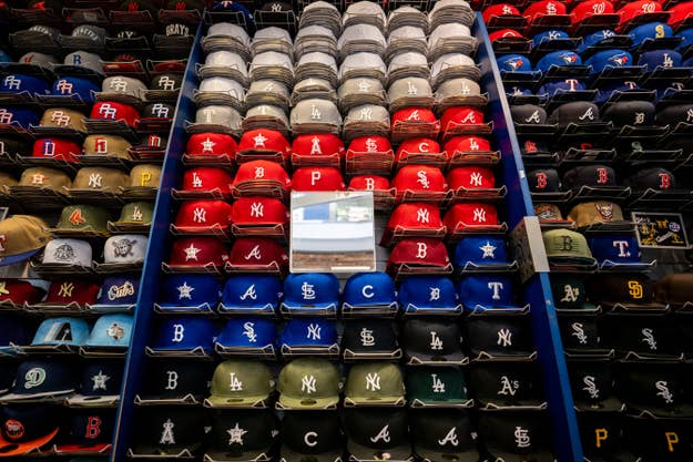 A store display shows rows of various baseball caps with team logos, including New York Yankees, Atlanta Braves, and others, arranged by color and team