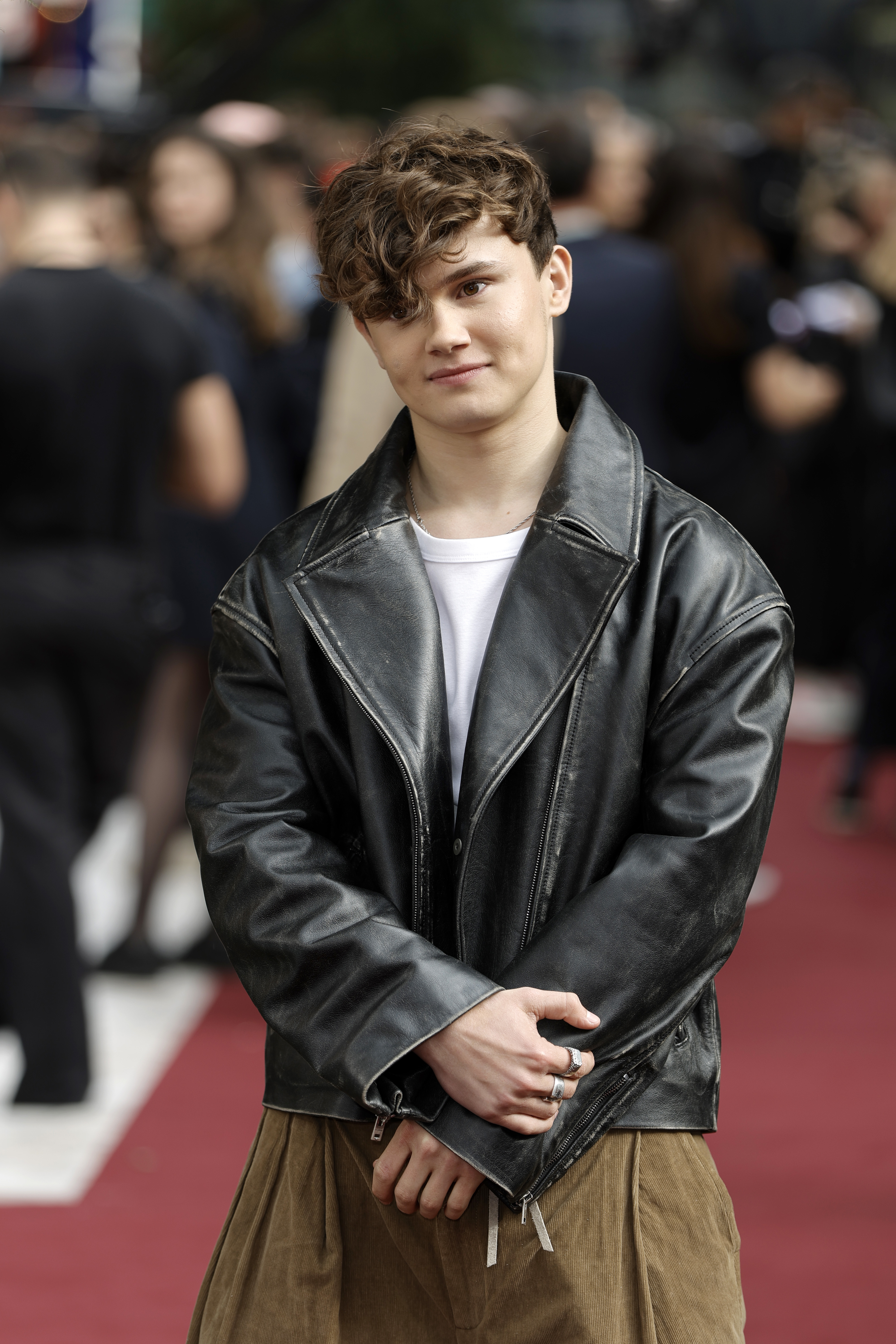 Will Tilston with wavy hair on a red carpet wearing a white T-shirt, oversized black leather jacket, and brown trousers