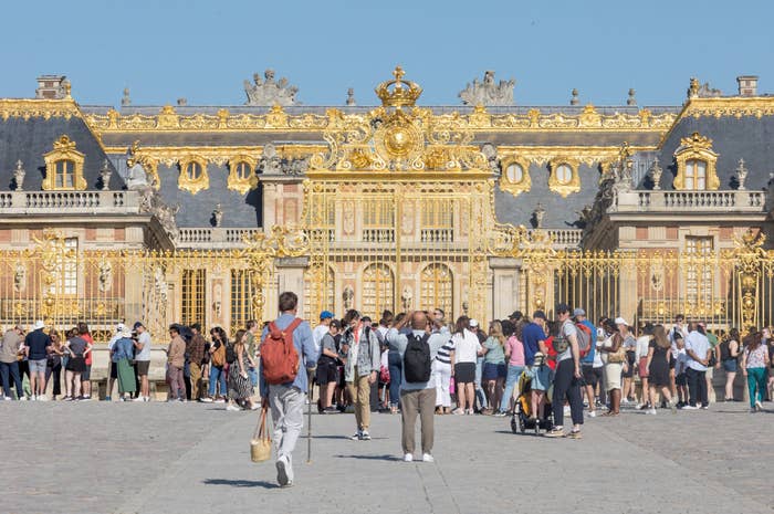 A large group of tourists stand in front of the ornate golden gates of the Palace of Versailles. They are taking photos and admiring the historic site