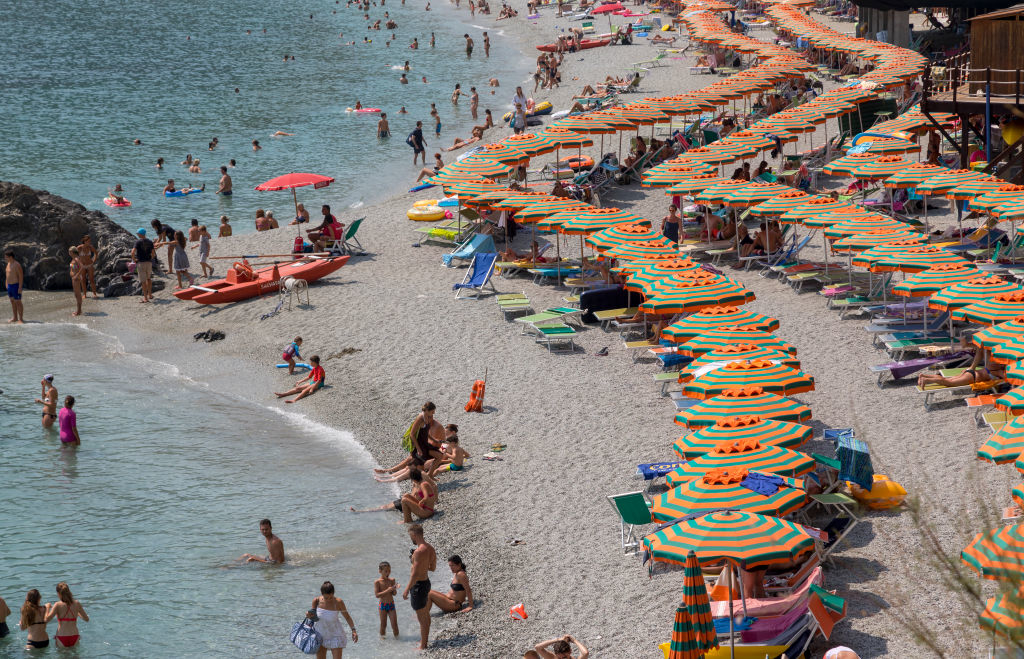 A crowded beach with many people sunbathing and swimming. Numerous colorful umbrellas in a row cover beach chairs along the shoreline
