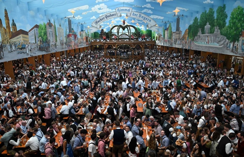 A large crowd is enjoying Oktoberfest inside a festively decorated beer hall, filled with people drinking, socializing, and wearing traditional Bavarian outfits