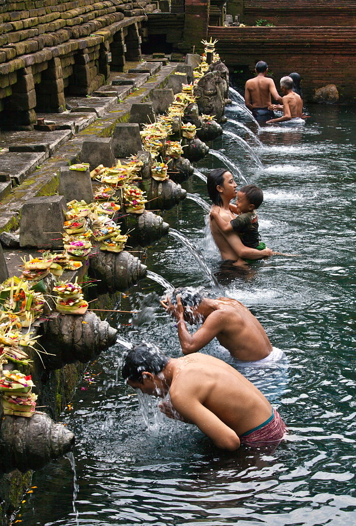 People bathing under fountains in a stone pool at a traditional outdoor purification ritual. Offerings are on the fountains
